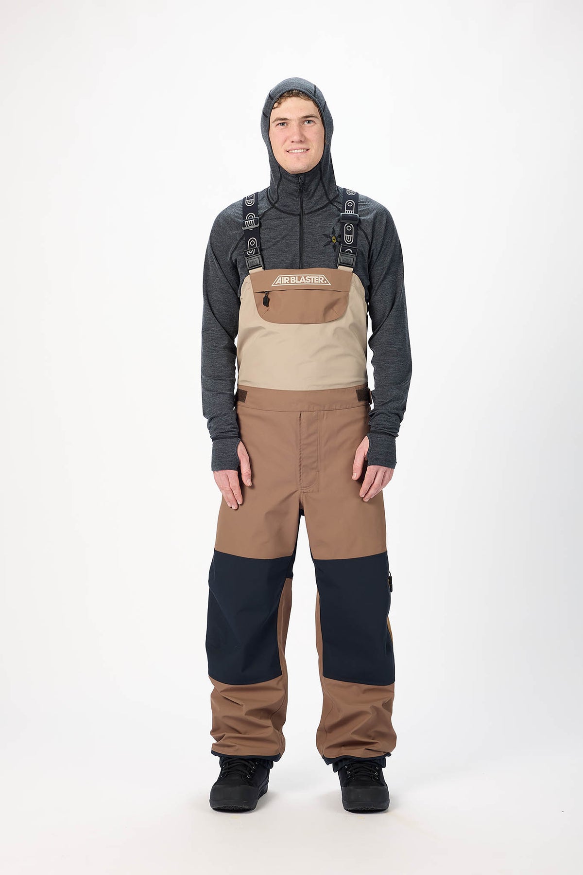 Person wearing a gray hoodie and tan overalls with black pants on a white background