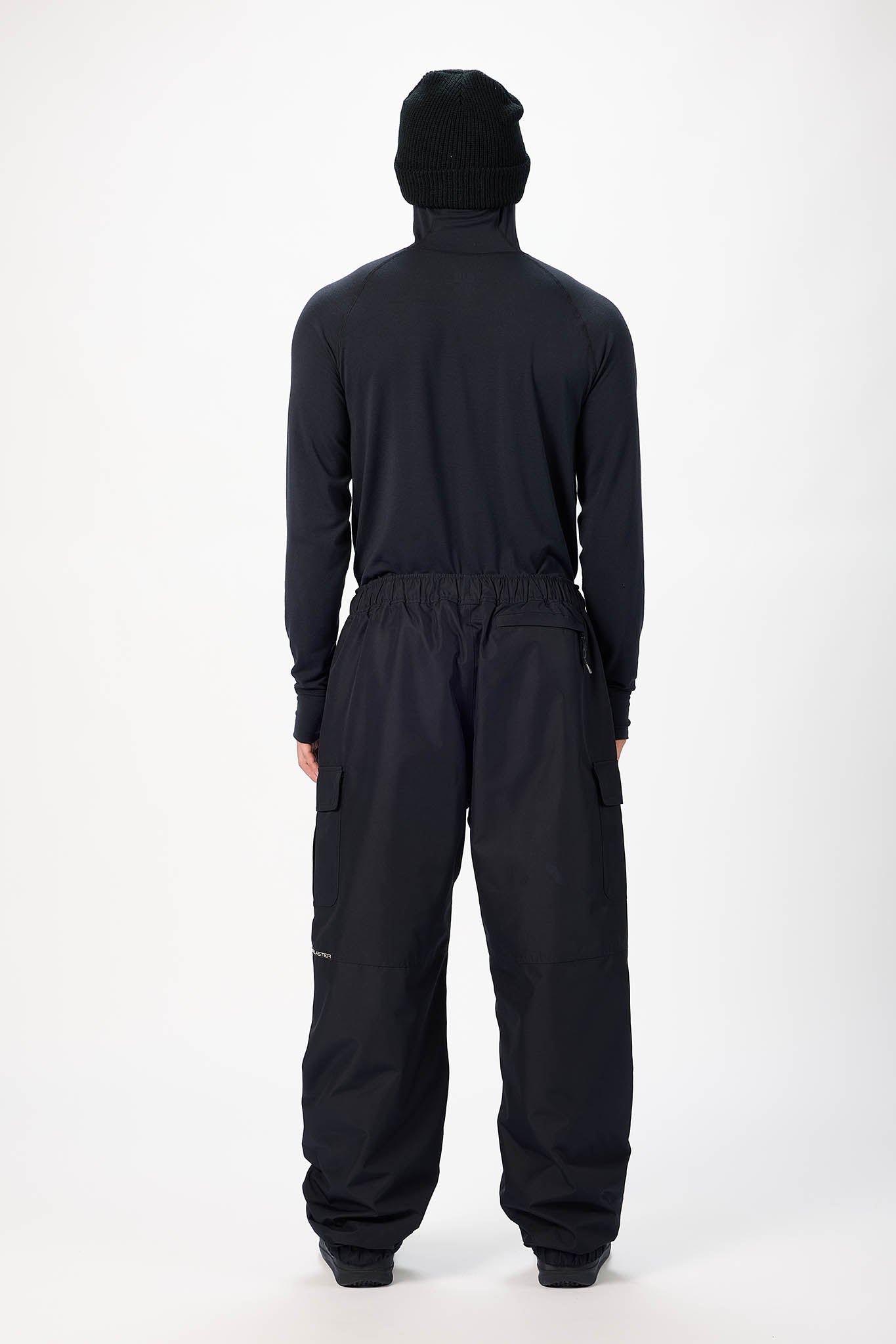 A person viewed from the back wearing a black hooded long-sleeve top and black wide-leg pants against a plain light gray studio background.