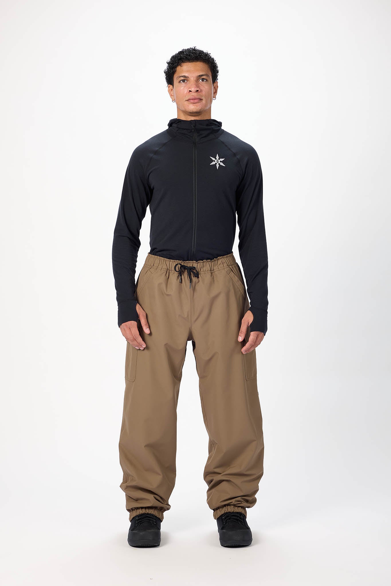 A man modeling a black long-sleeve zip-up hoodie with a white geometric logo on the left chest, paired with brown drawstring pants and black shoes, against a plain white studio background.