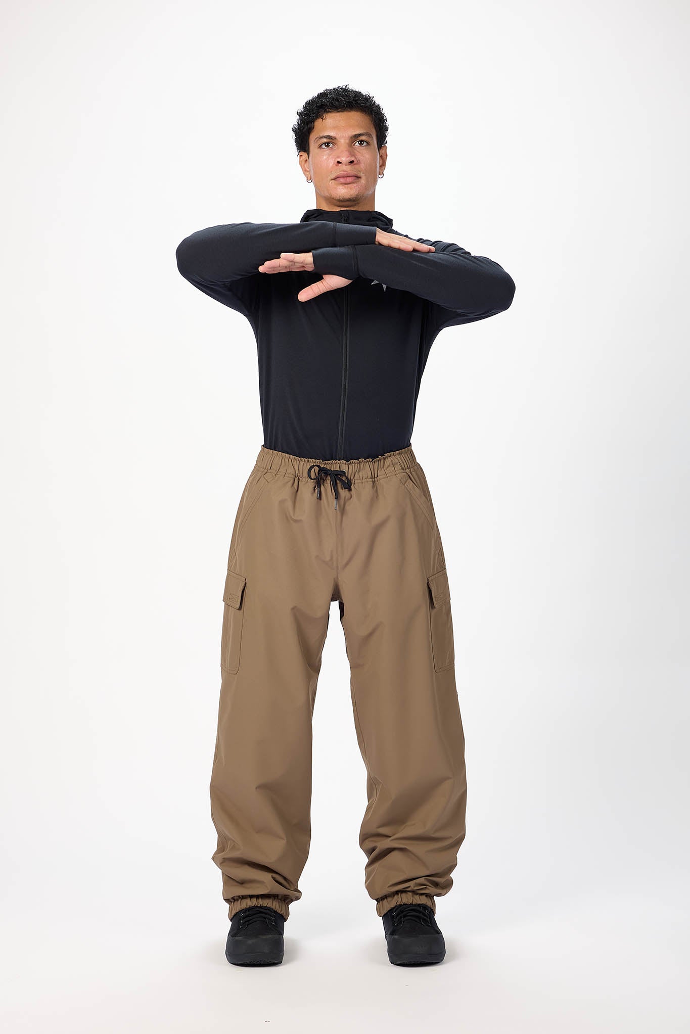A man modeling a black long-sleeve zip-up top and brown drawstring cargo pants against a plain white studio background.