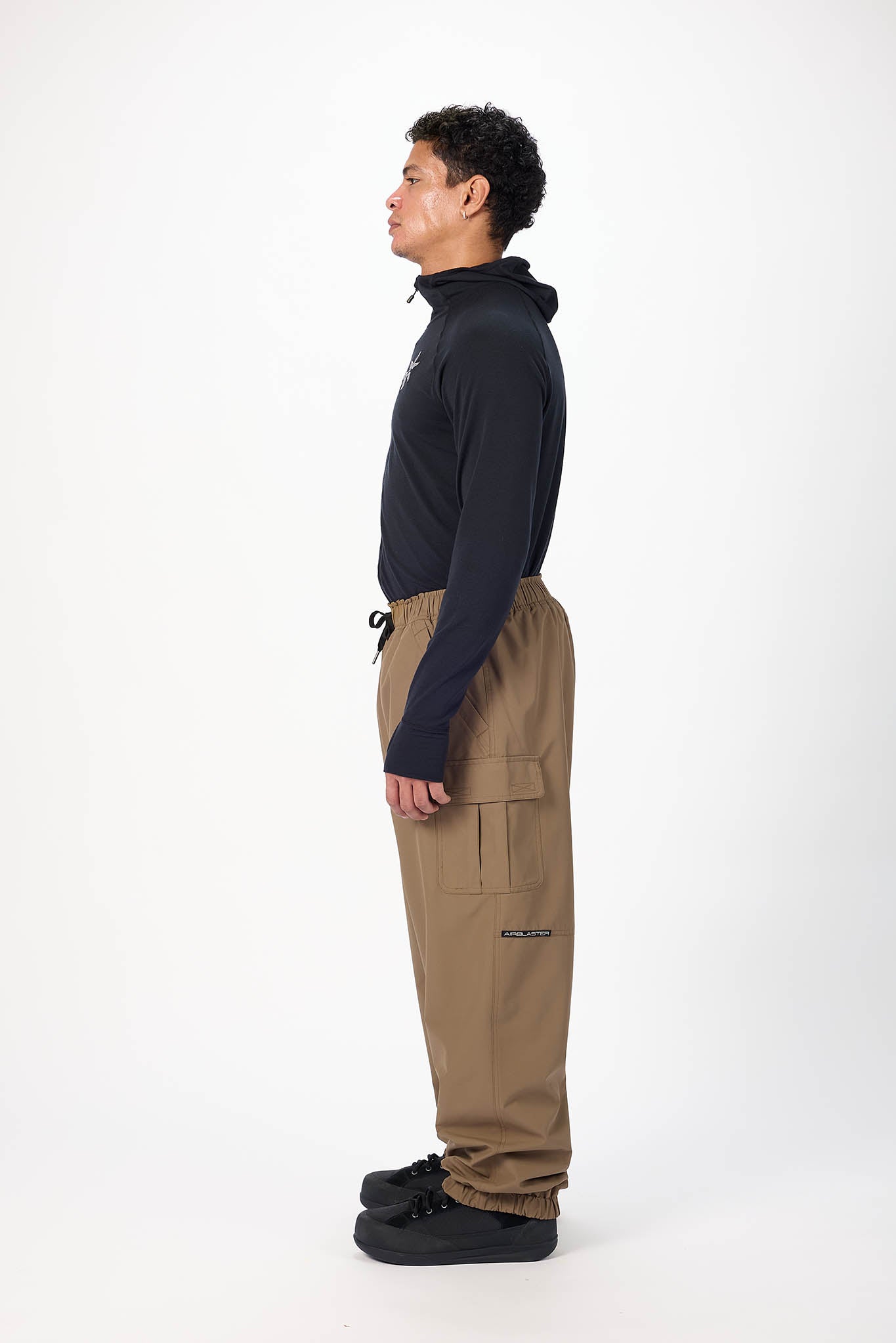 A man shown in profile wearing a black long-sleeve top and brown cargo pants against a plain white studio background.