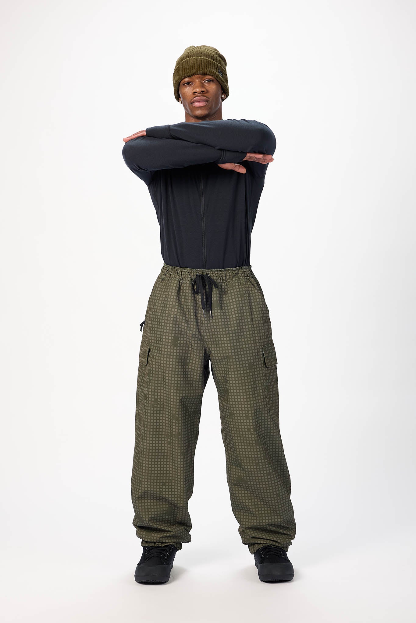 A model stands against a white studio background wearing an olive green knit beanie, a black long-sleeve shirt, olive green checkered cargo pants with a black drawstring, and black sneakers.