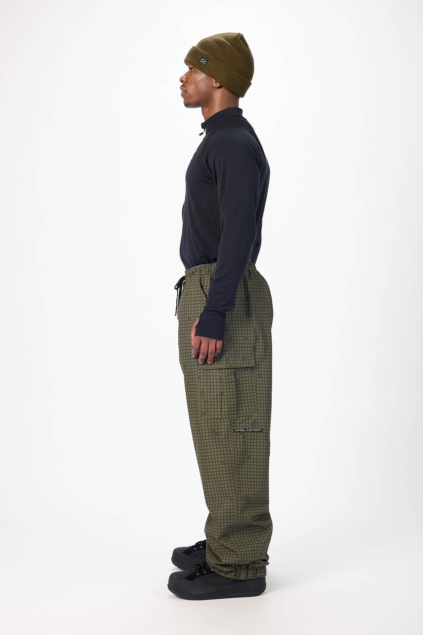 A man shown in profile wearing a dark navy long-sleeve top, olive green patterned pants, a matching olive green beanie, and black shoes, standing against a plain white studio background.