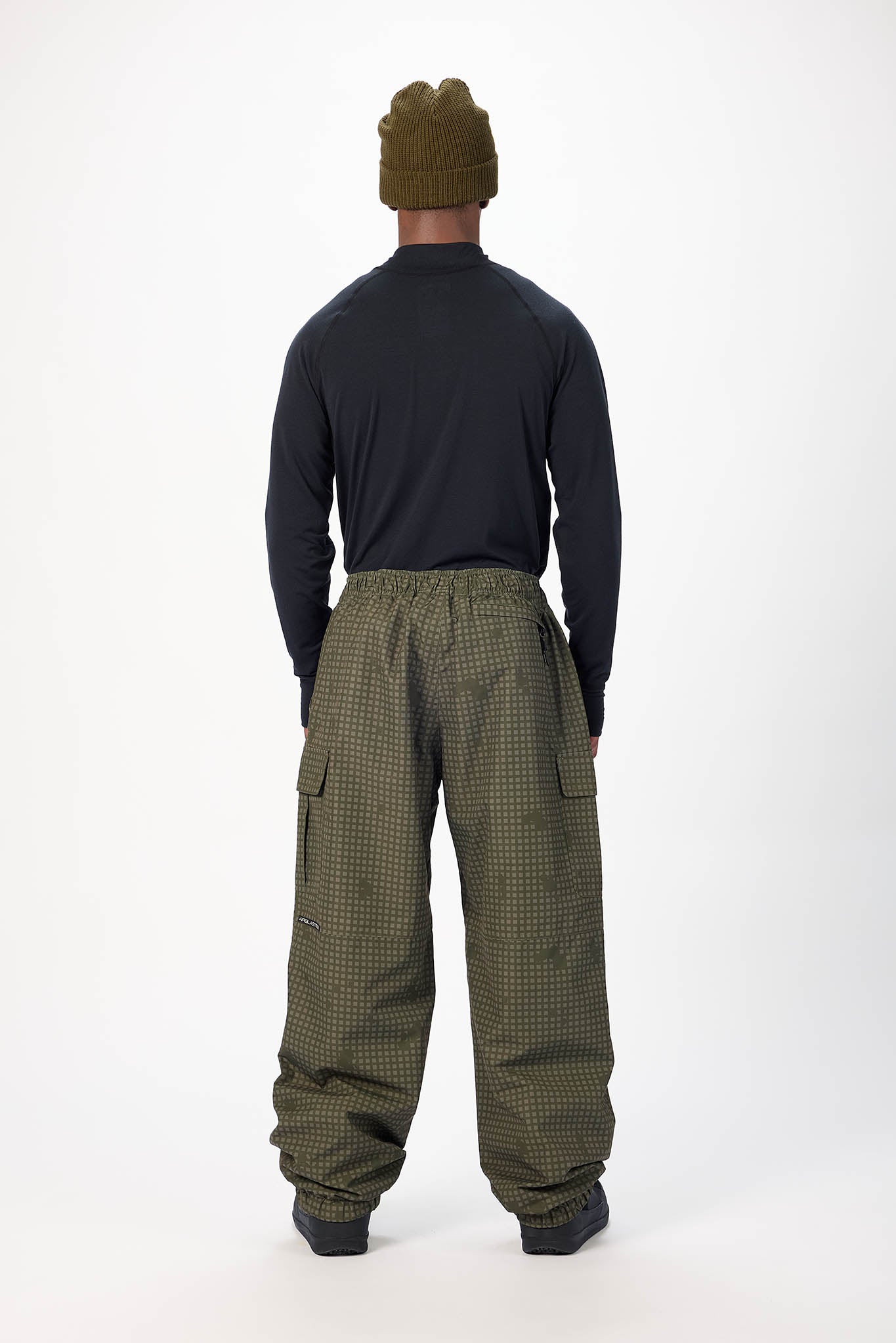 A person shown from the back wearing wide-leg olive green patterned pants, a black long-sleeve top, and an olive green beanie against a plain white studio background.