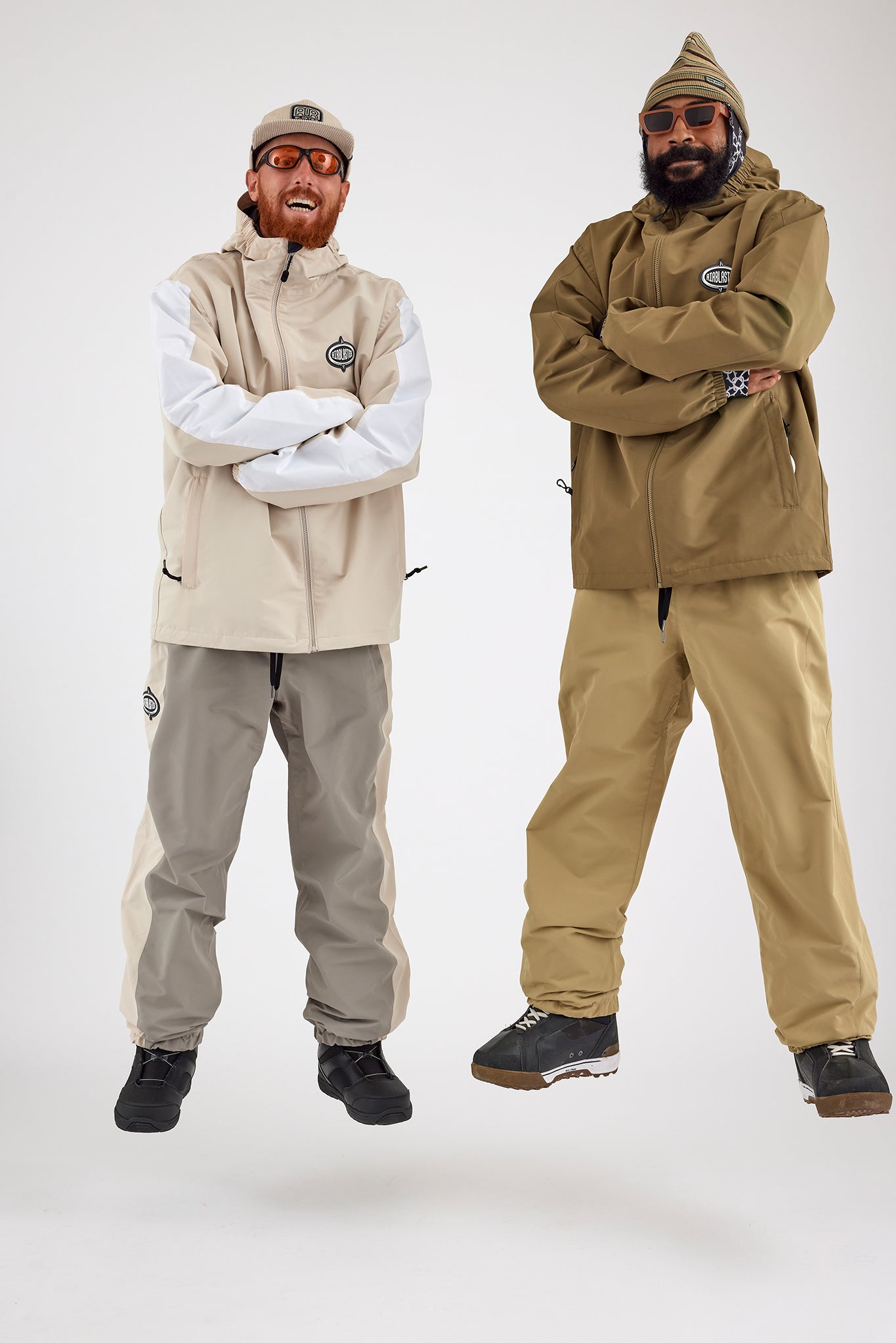 Two individuals, both dressed in winter sportswear, are playfully jumping with arms crossed. One wears a beige and white waterproof jacket with a cap and sunglasses, while the other sports eco-friendly Airblaster Access Jacket in olive-green with a knitted hat and sunglasses. The background is white.