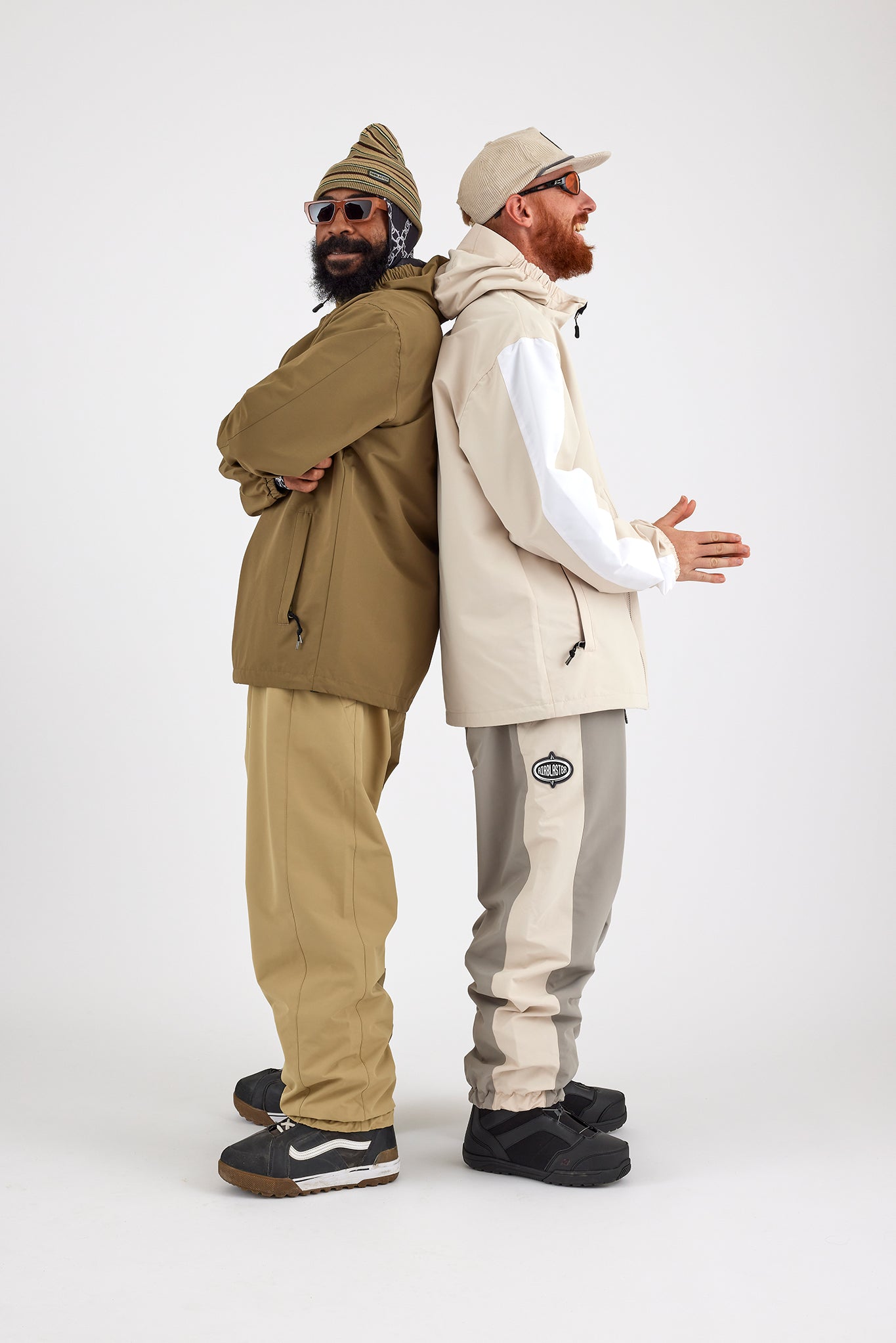 Two men stand back-to-back against a white background. The man on the left wears an olive green outfit with a beanie, sunglasses, and black shoes. The man on the right, in a beige and white jacket, light grey Airblaster Access Pant with 10K waterproofing, beige cap, and black shoes, smiles with his hands up.