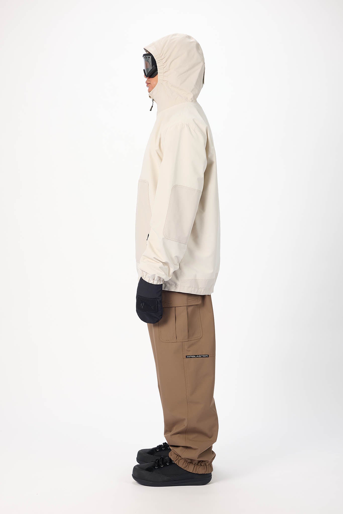 A side-profile studio shot of a person wearing a light beige hooded jacket, brown pants, black gloves, and black shoes against a plain white background.