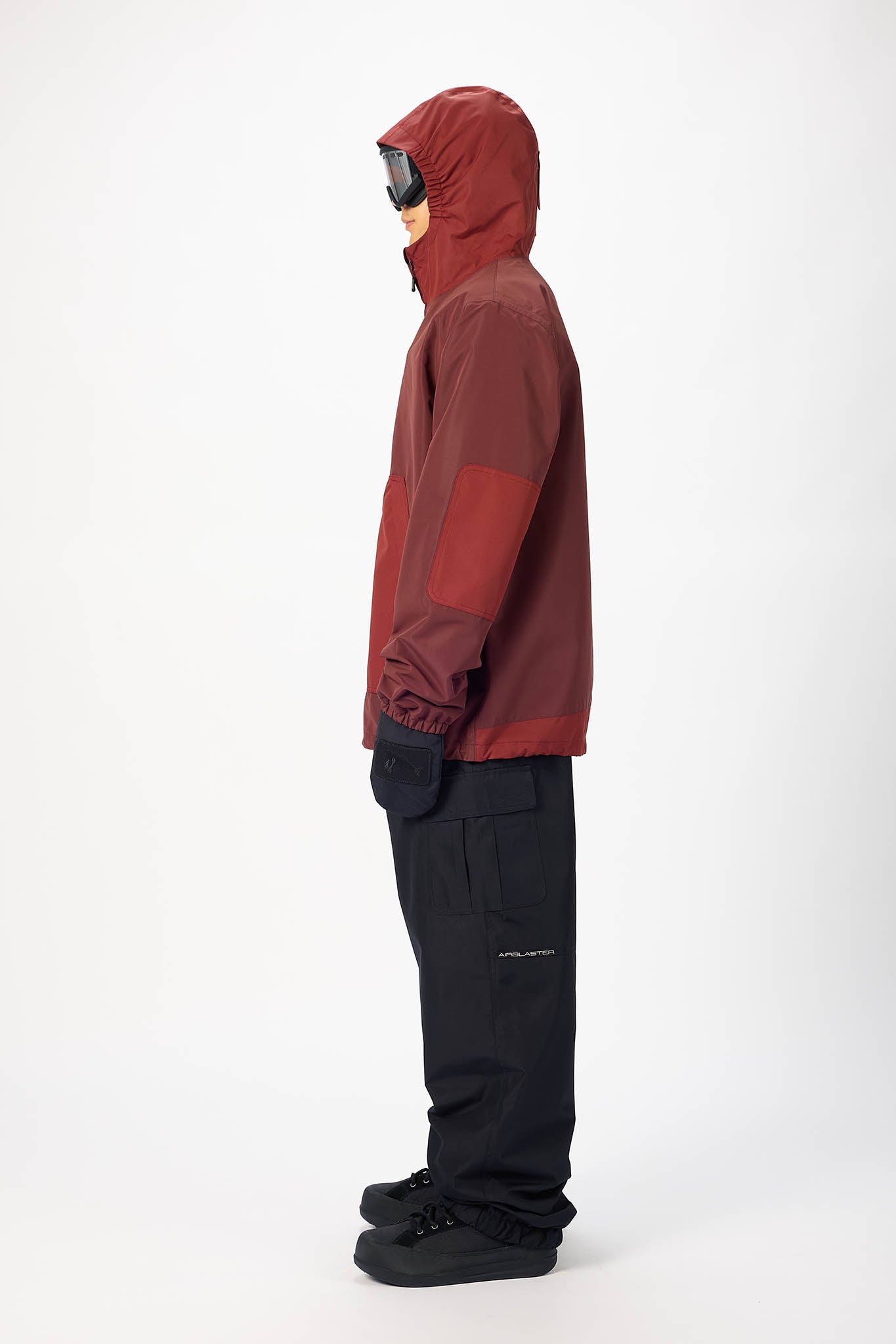 A side-profile studio shot of a model wearing a dark red hooded technical jacket with black snow pants and black ski boots, set against a plain white background.
