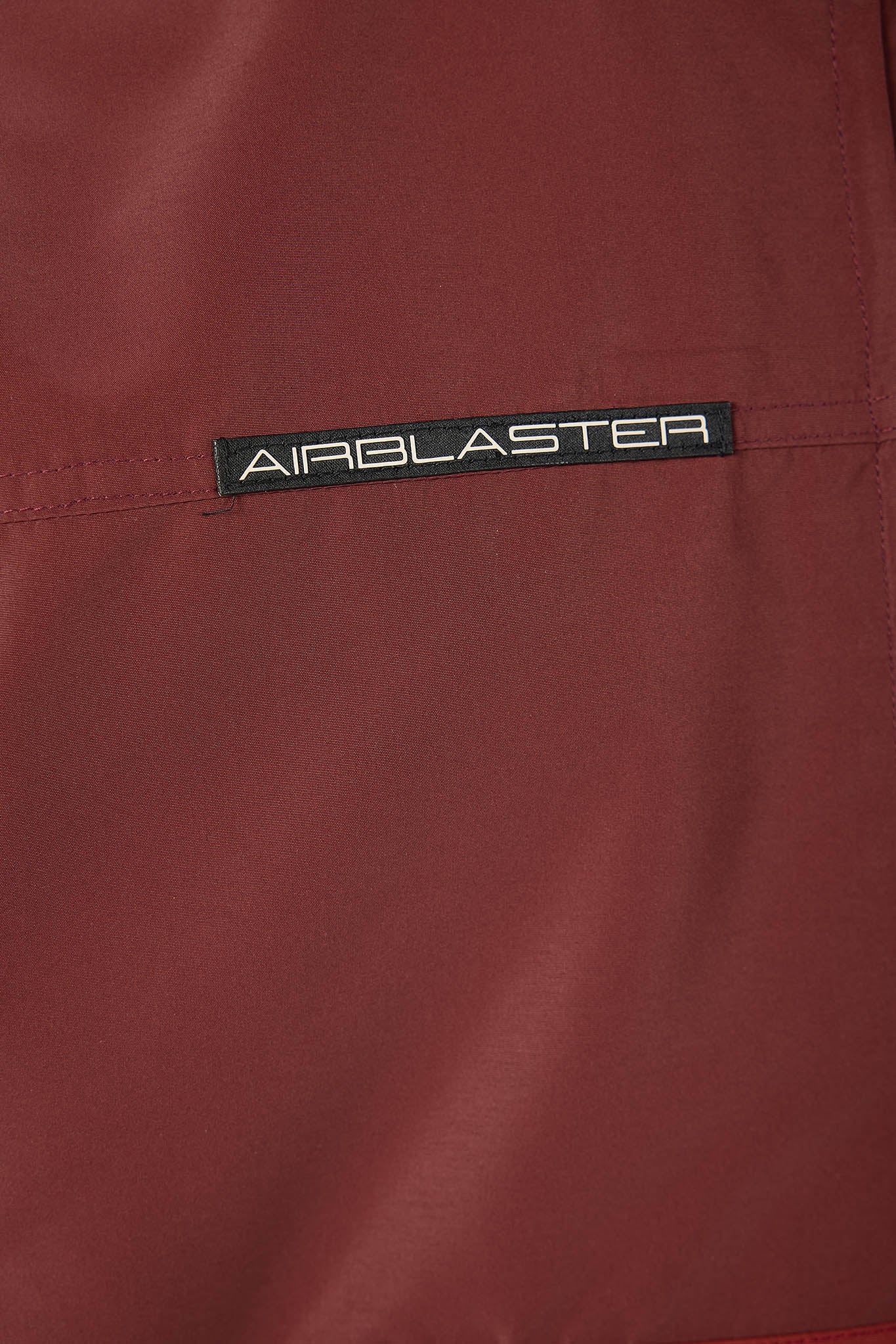Close-up of a dark red fabric surface with a black rectangular AIRBLASTER logo patch stitched horizontally across the center.