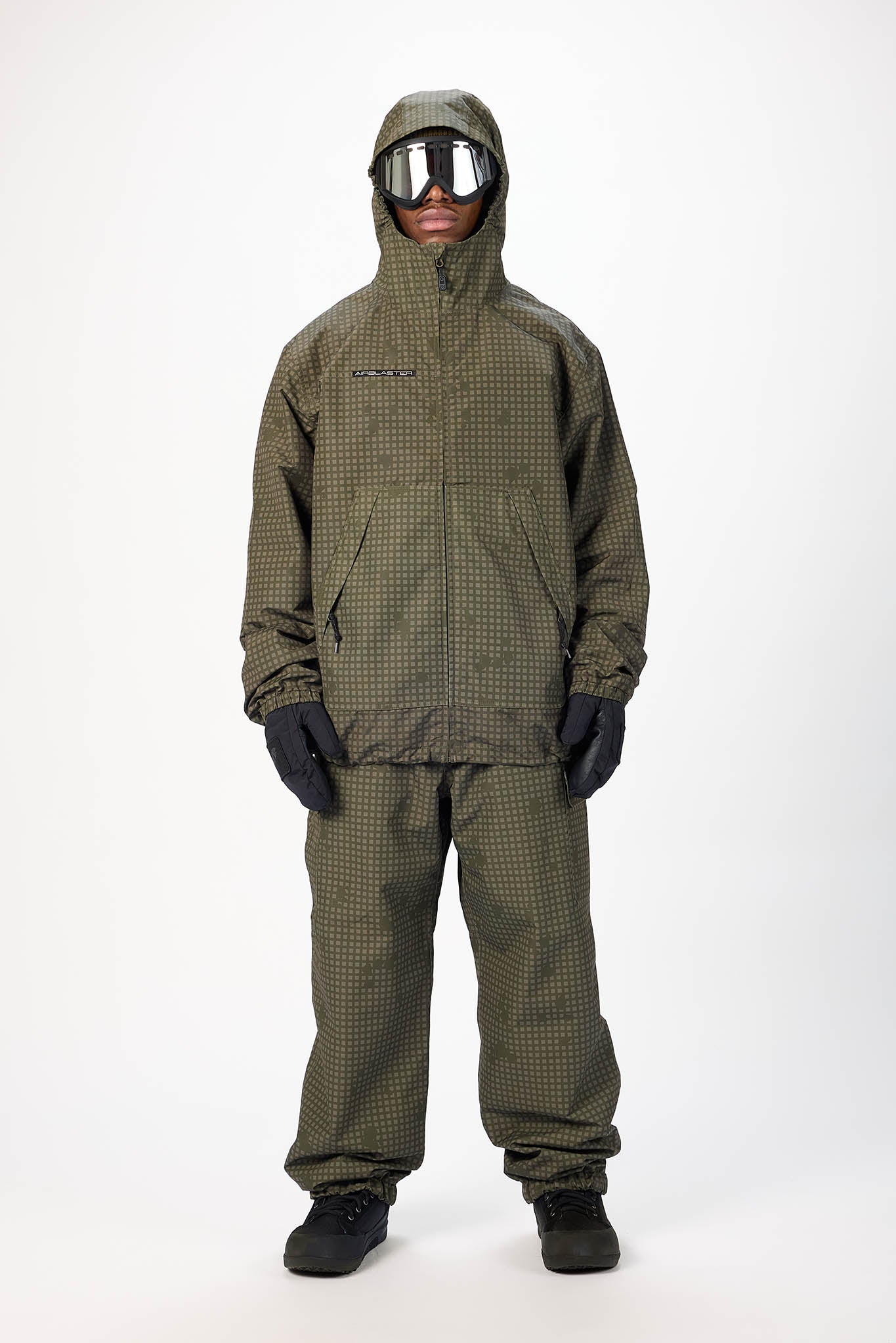A model wearing a hooded snowsuit in an olive green and black checkered pattern, complete with black gloves, black snow boots, and reflective goggles, standing against a plain white studio background.
