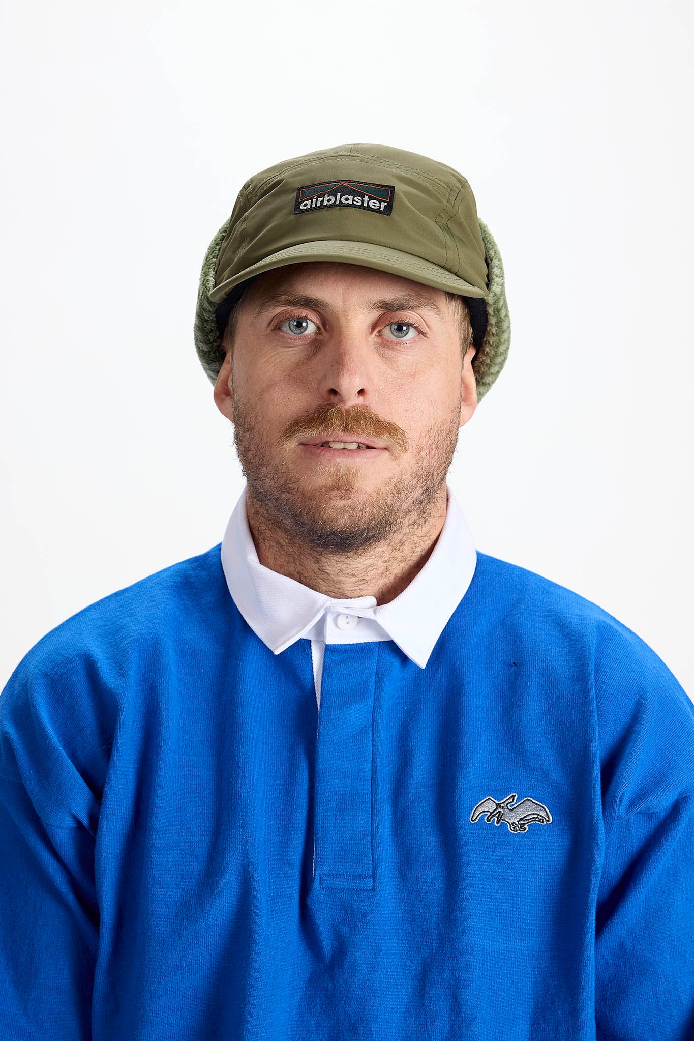 A man wearing an olive green Airblastes cap, a blue rugby-style polo shirt with a white collar, and a green knit beanie, photographed against a plain white studio background.