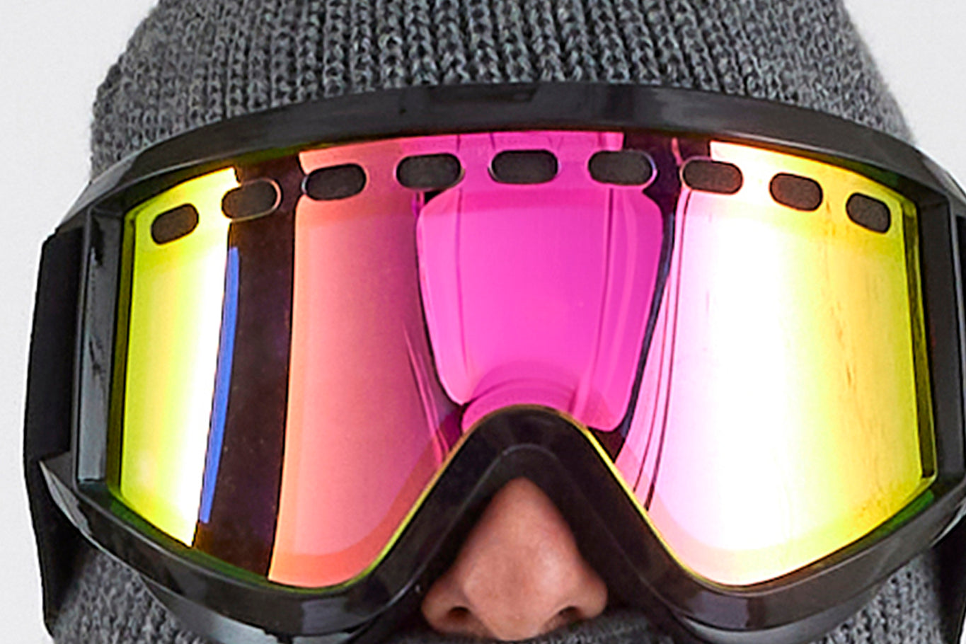 A person wearing a gray knit hat and reflective ski goggles with yellow and pink tints. The Airblaster Air Goggle, designed with UV protection, has a black frame and large lenses that cover the person's eyes and nose for enhanced safety.