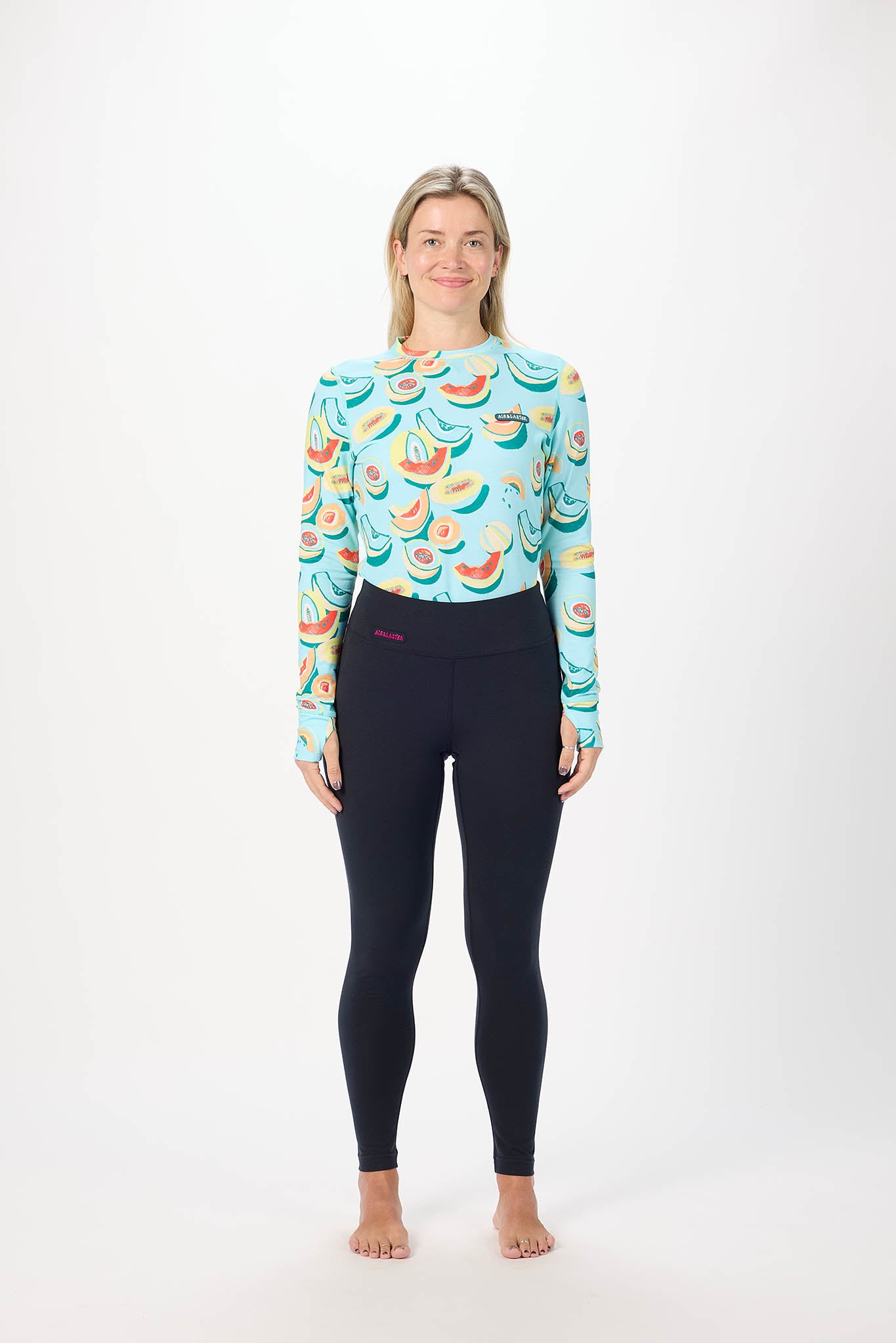 A woman stands against a plain white studio background wearing a long-sleeve athletic top with a colorful watermelon and banana print on a light blue base, paired with black leggings.