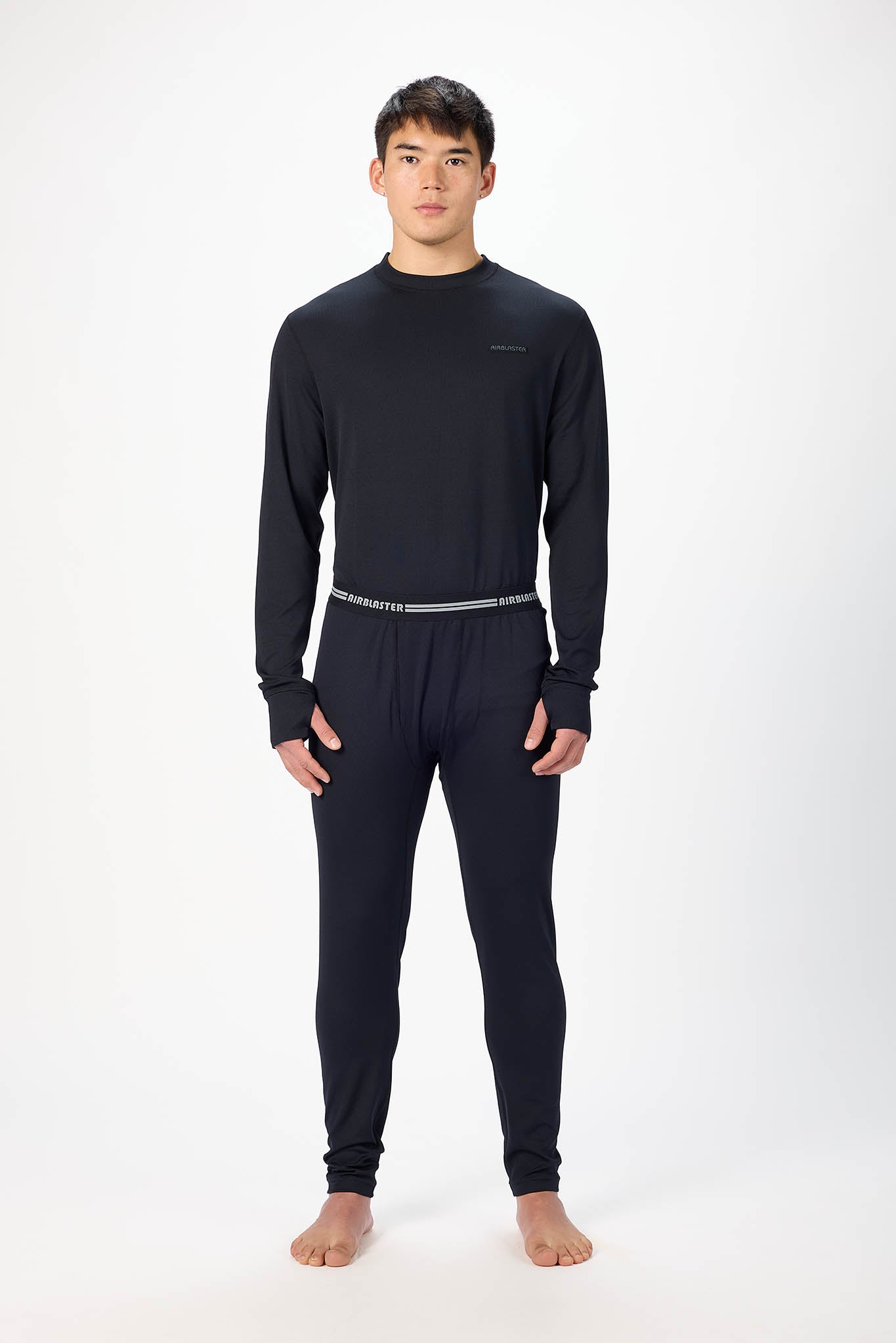 A man modeling a black long-sleeve crewneck base layer and matching black pants with a thin silver waistband, standing against a plain white studio background.