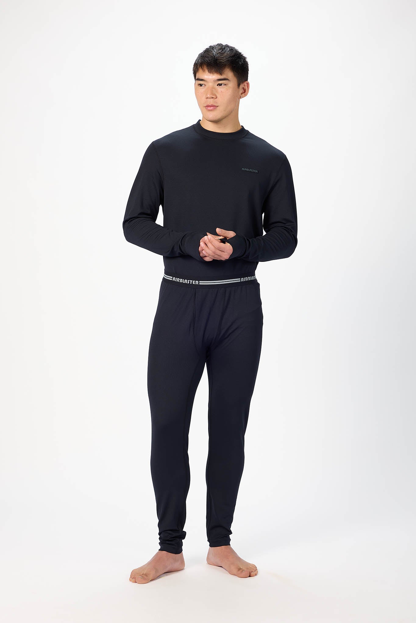 A man modeling a black long-sleeve crewneck base layer and matching black pants with a thin silver waistband, standing against a plain white studio background.