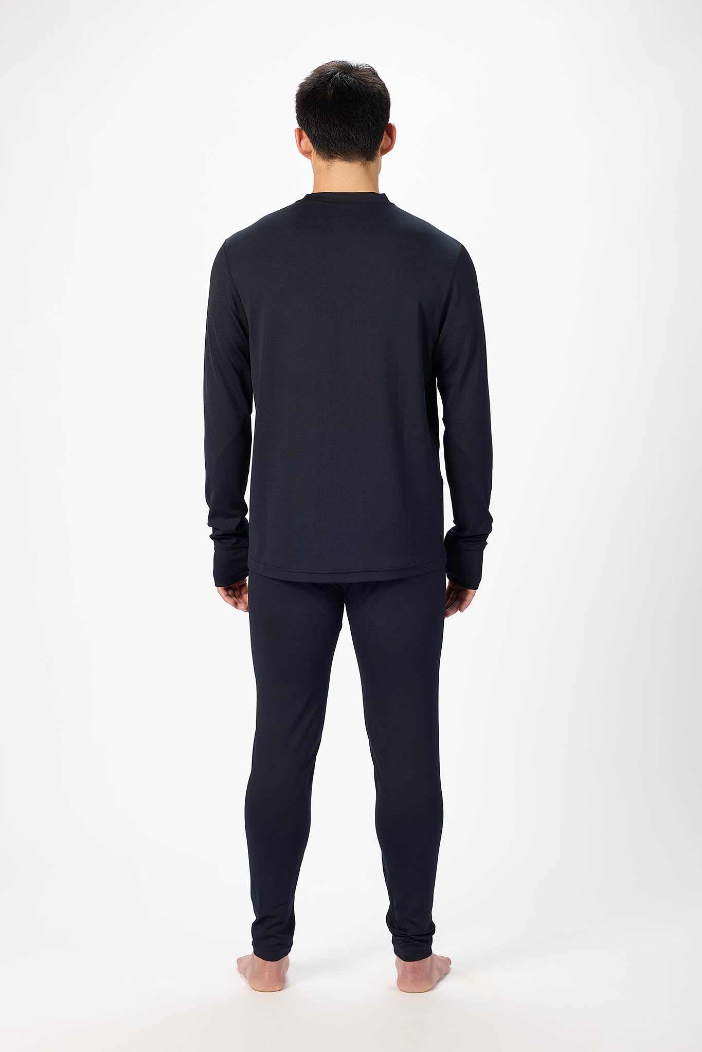 A man viewed from behind wearing a dark navy long-sleeve top and matching navy pants, standing barefoot against a plain white studio background.
