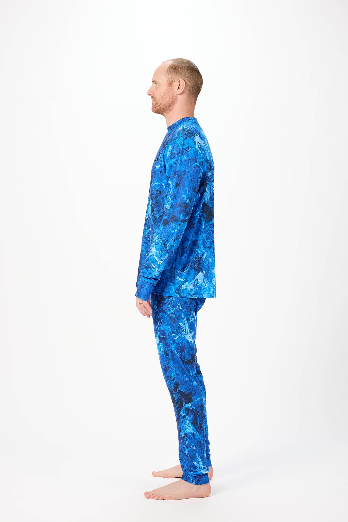 A man shown in profile wearing a matching long-sleeve top and pants set with an abstract blue and black pattern, standing barefoot against a plain white studio background.