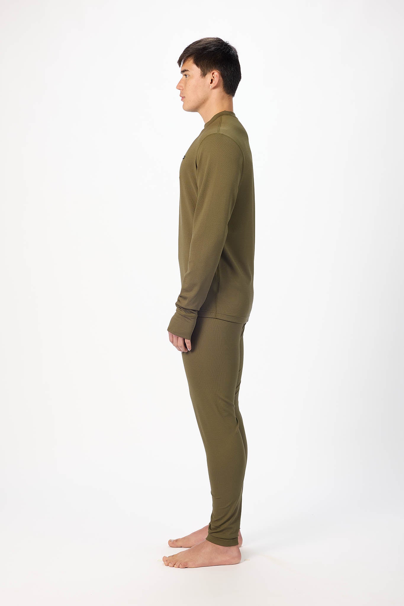 A man in profile wearing an olive green long-sleeve top and matching pants, standing barefoot against a plain white studio background.