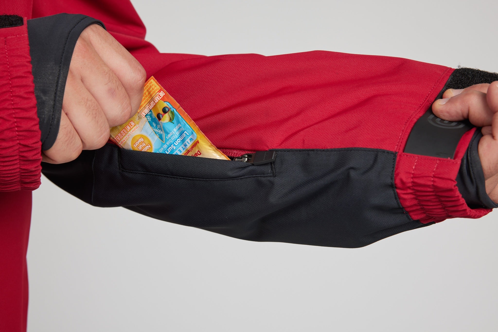Close-up of a person's hands demonstrating a zippered arm pocket on a red and black jacket, with a small snack packet tucked into the pocket.