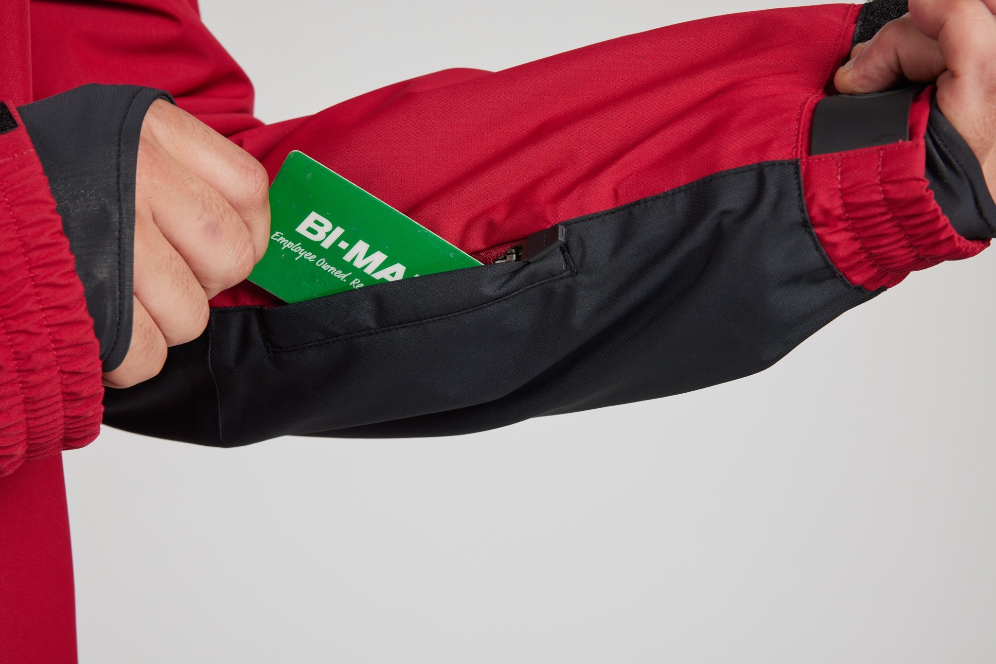 Close-up of a person wearing a red and black ski jacket, pulling the sleeve open to show a green Bi-Mat thermal pocket insert.