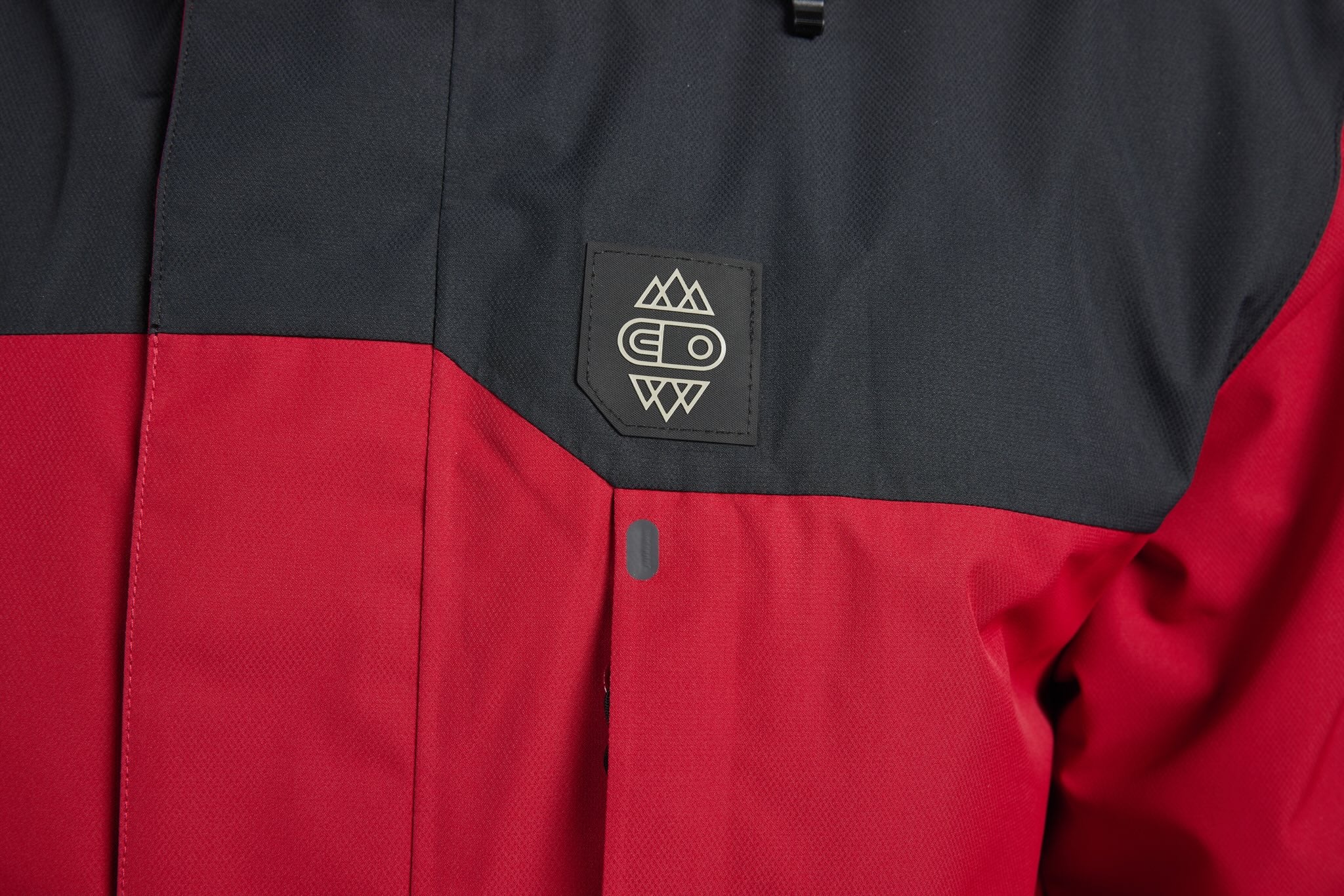 Close-up of a colorblock jacket with a dark gray upper section and a red lower section, featuring a black rectangular patch on the chest with a white mountain