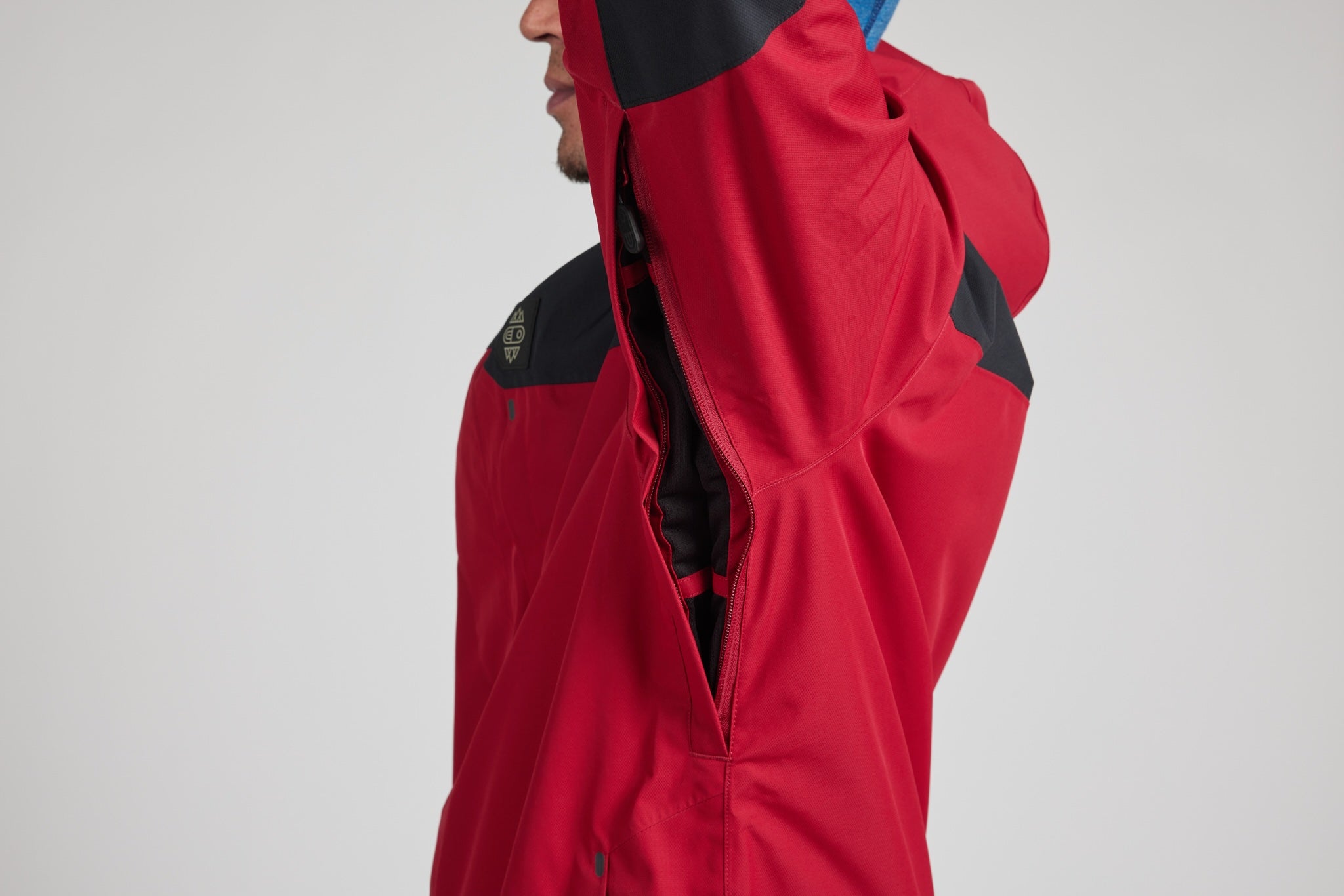 A close-up side profile of a person wearing a red and black technical outdoor jacket, with the hood up and a zipper opening, against a light gray studio background.