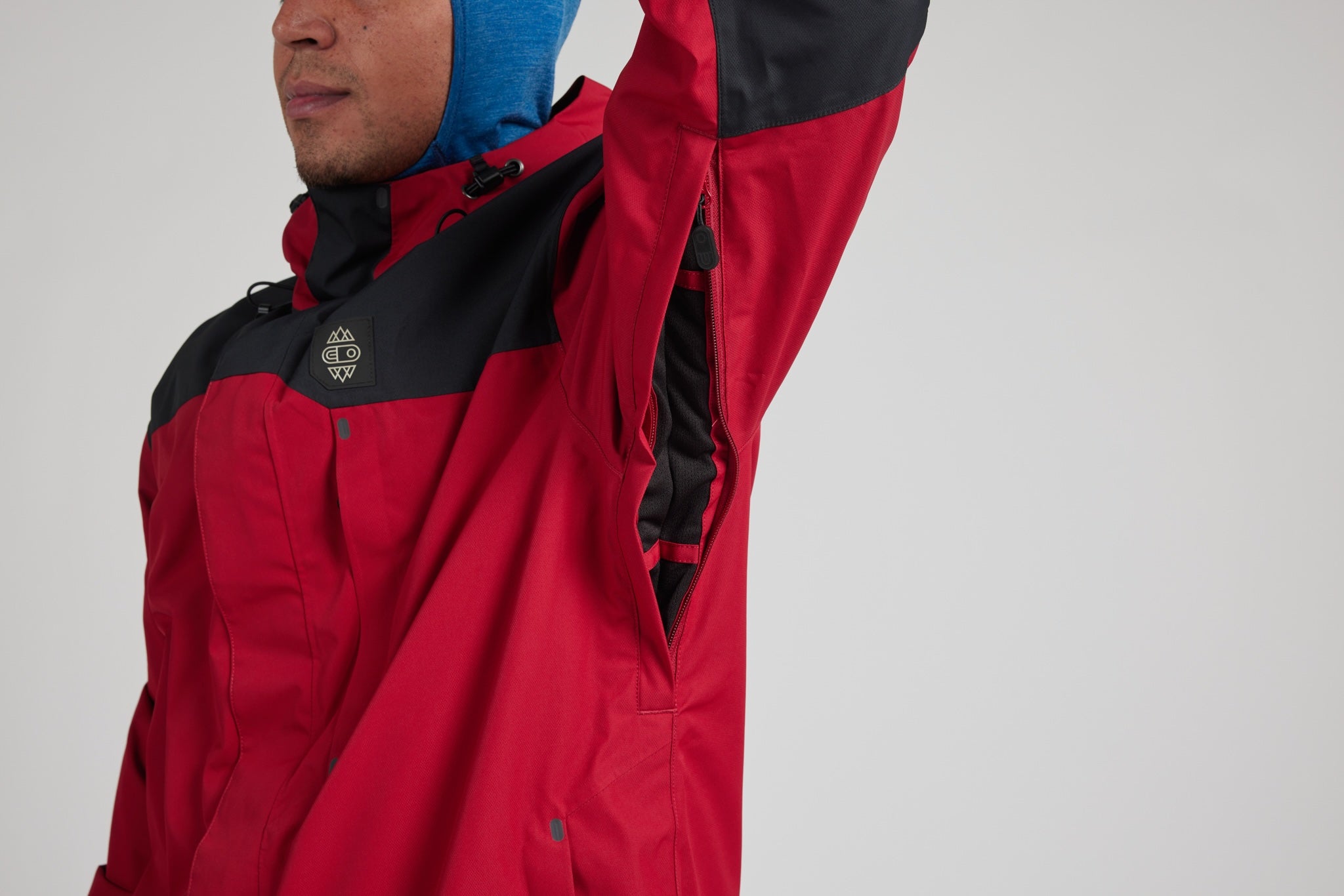 A close-up of a person wearing a red and black ski jacket, with one arm raised, against a plain light gray studio background.