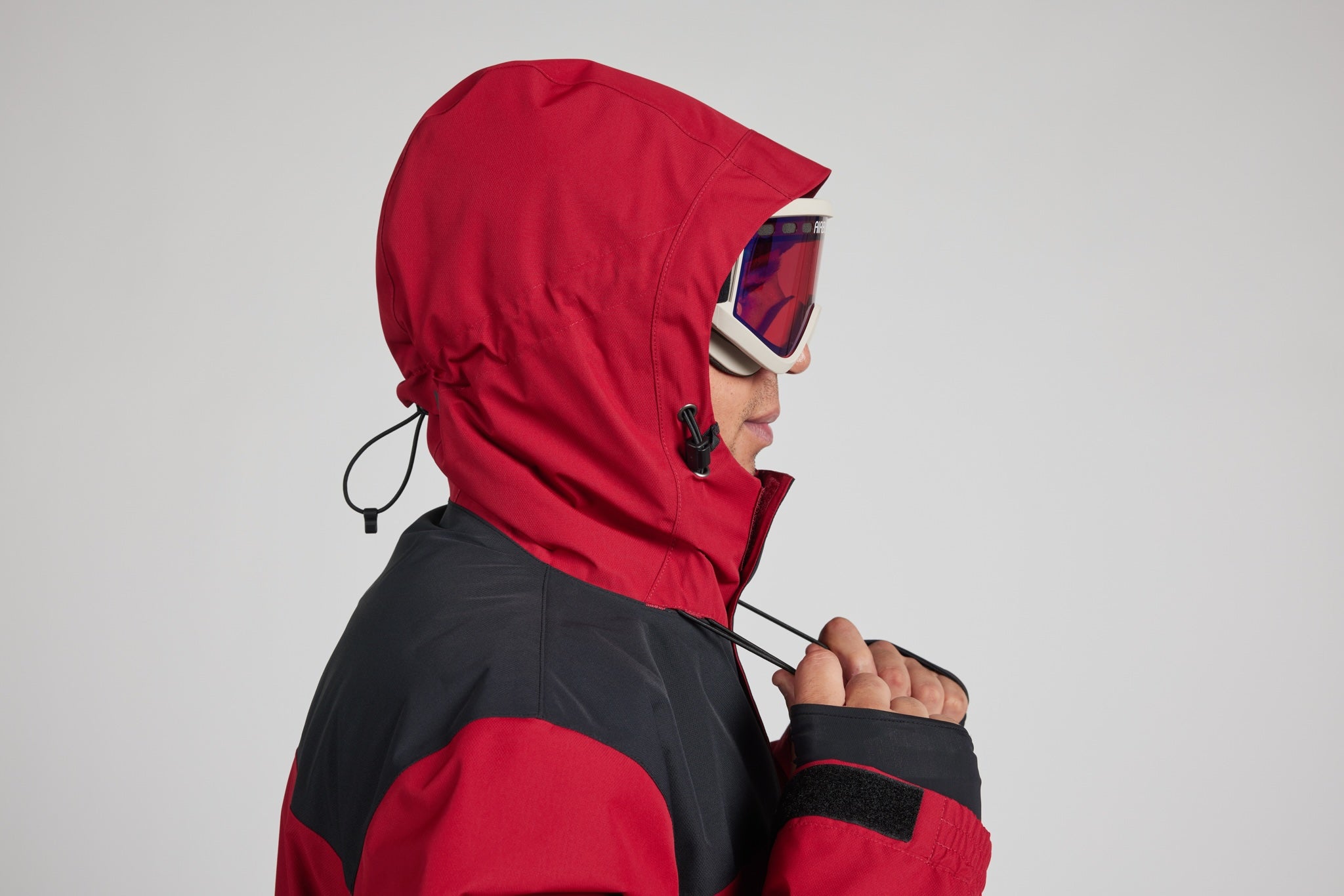 A person wearing a red and black ski jacket with the hood up, shown in profile against a light gray studio background. The jacket has a red hood and upper chest, with a black lower body panel.