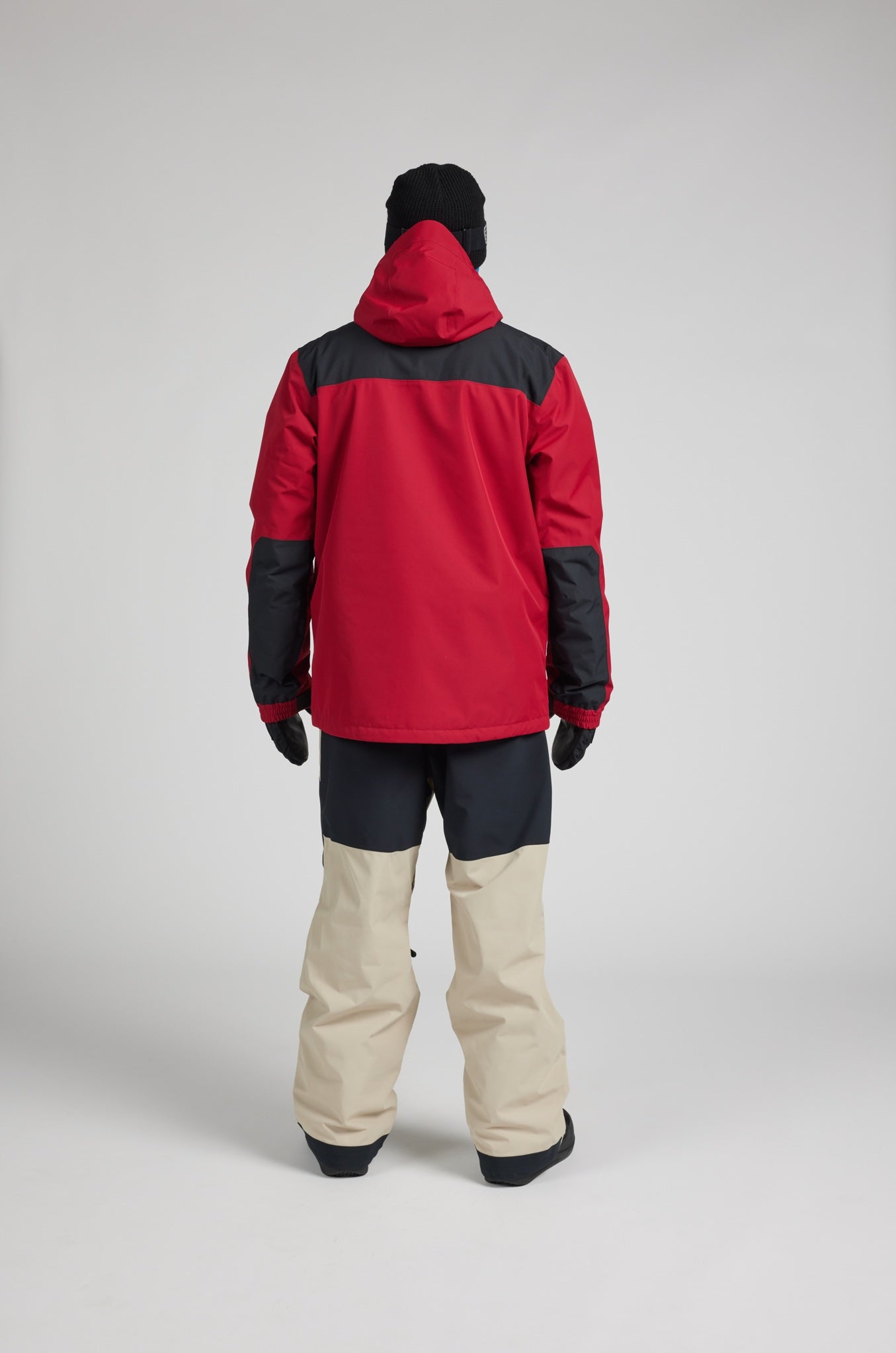 A model viewed from the back wearing a red and black color-blocked hooded jacket, beige and black snow pants, black gloves, and a black beanie, set against a plain light gray studio background.
