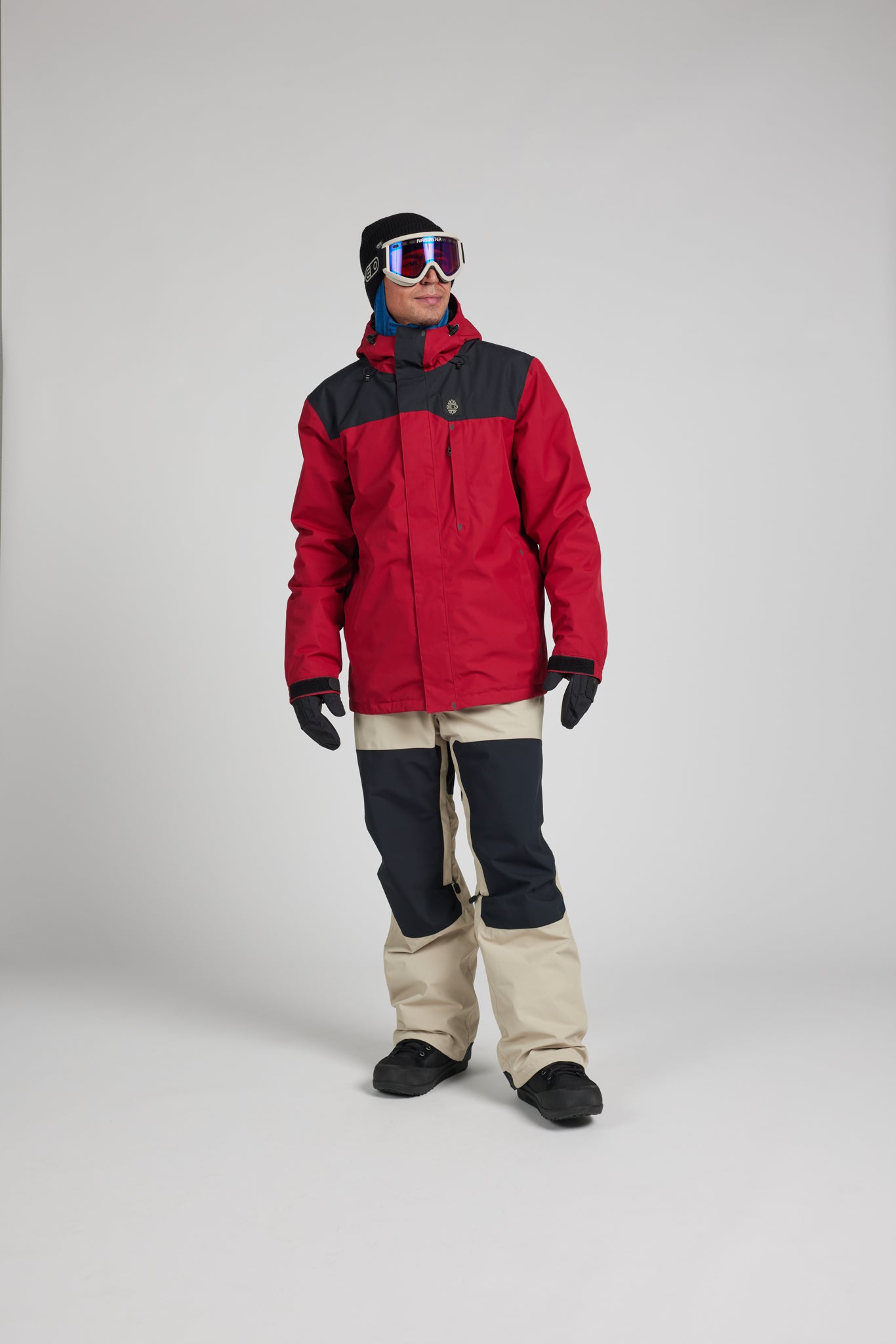 A model wearing a red and black ski jacket, beige and black snow pants, black gloves, and ski goggles, standing against a light gray studio background.