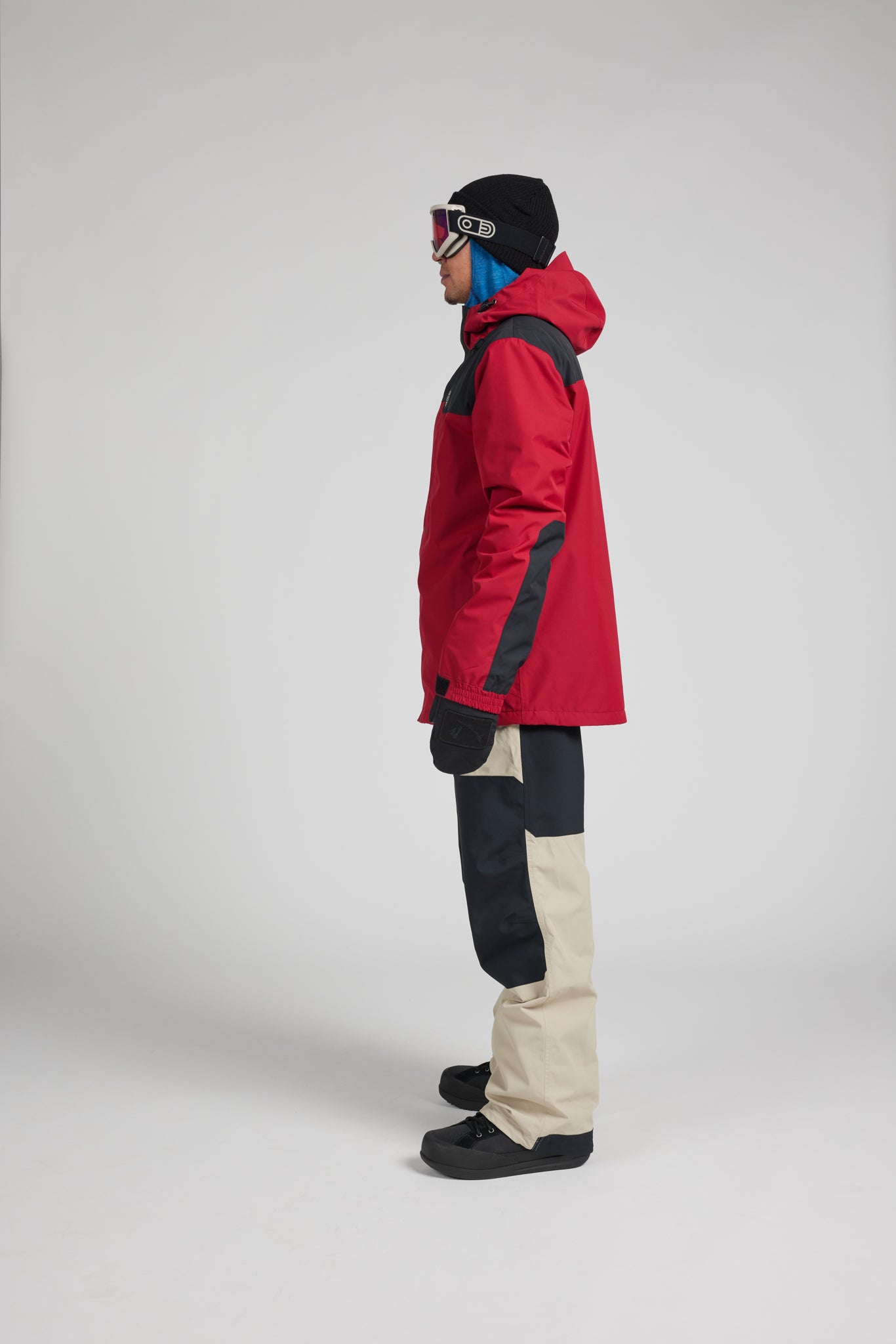 A side-profile studio shot of a model wearing a red and black ski jacket, beige and black color-blocked snow pants, a black beanie, and ski goggles against a light gray background.