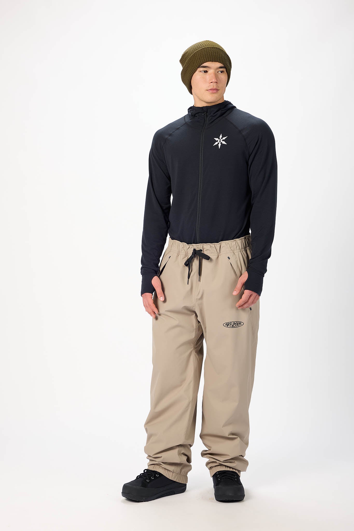 Person wearing a black long-sleeve shirt with a logo, beige pants, and a green beanie on a white background