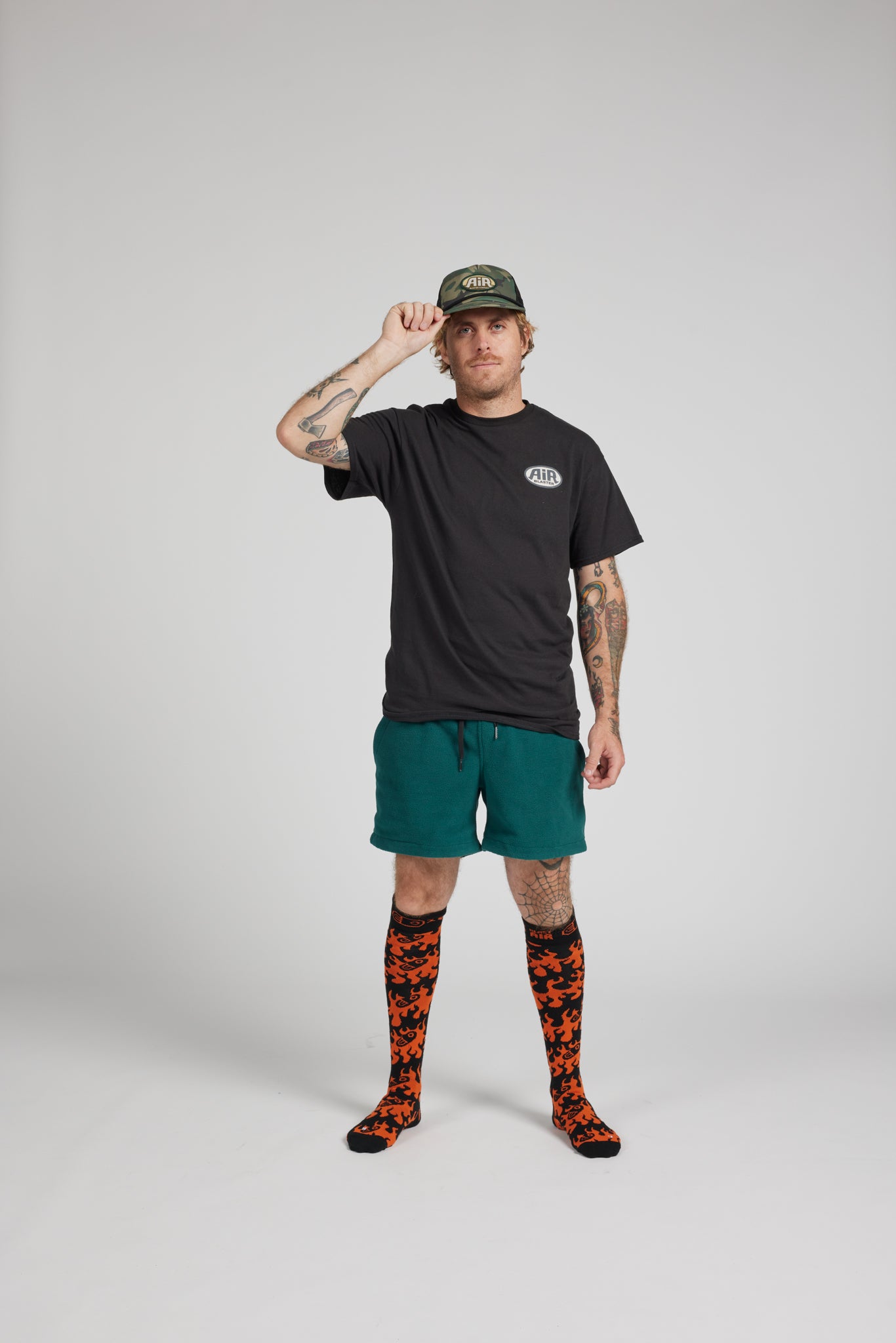 A man modeling a black t-shirt, green shorts, and orange-and-black patterned socks against a light gray studio background.