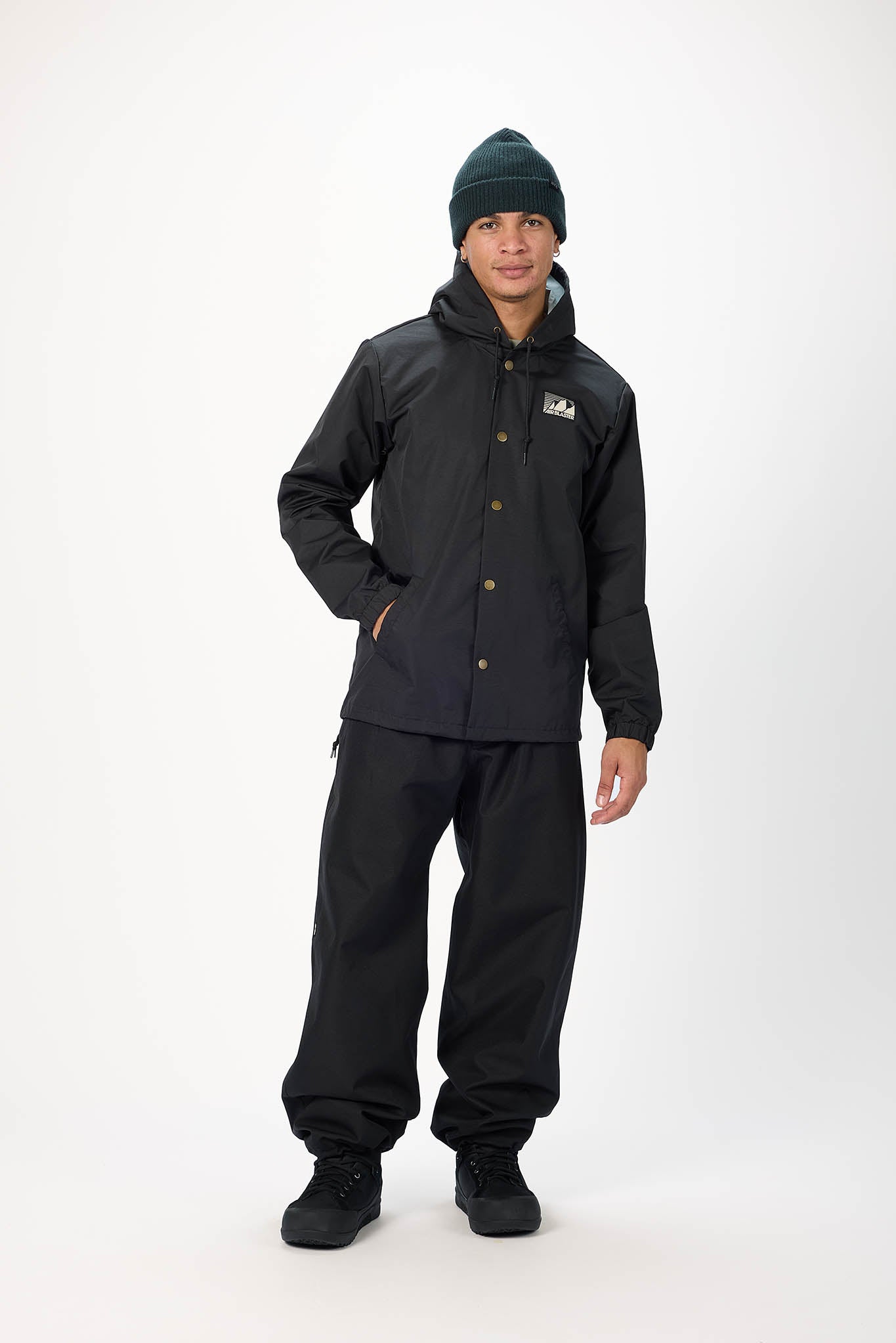A model wearing a black hooded jacket with brown buttons, black pants, and a dark green beanie, standing against a white studio background.