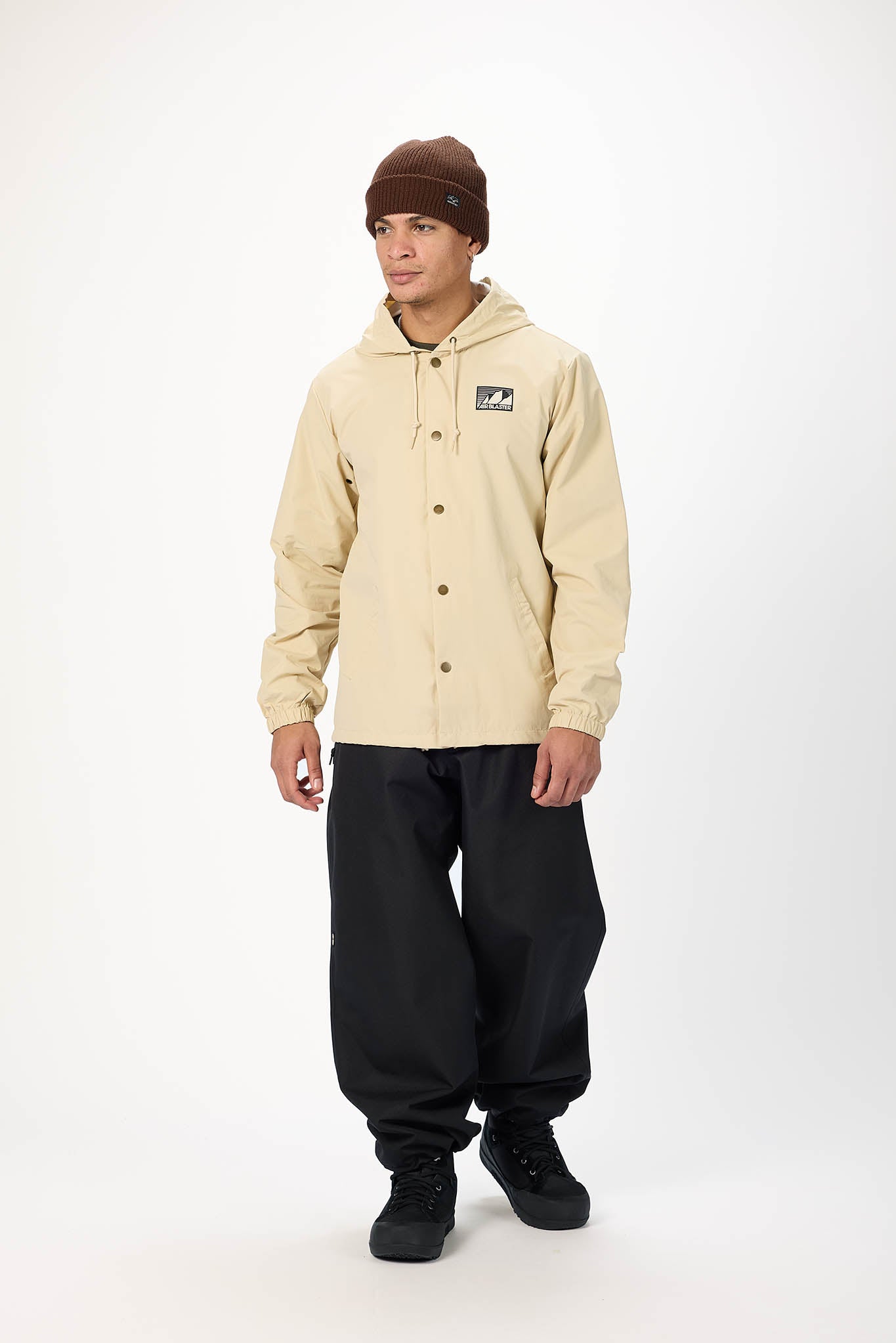 A model wearing a beige hooded jacket with a button front, black baggy pants, black boots, and a brown beanie, standing against a white studio background.