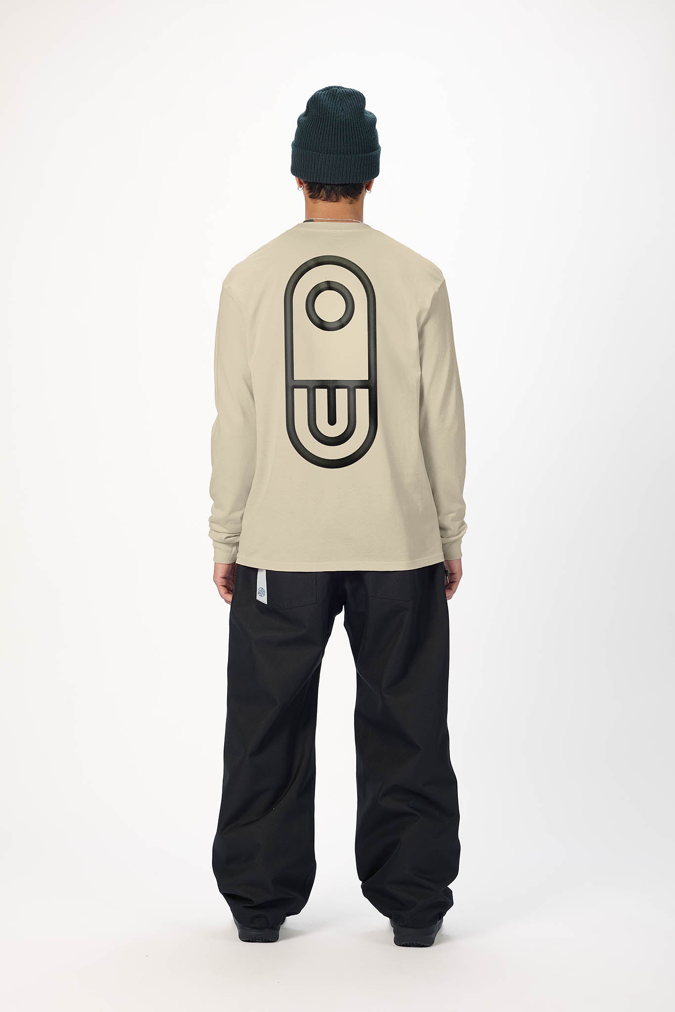 A person shown from the back wearing a light beige long-sleeve shirt with a large black graphic on the back, black pants, and a dark green beanie, against a plain white studio background.