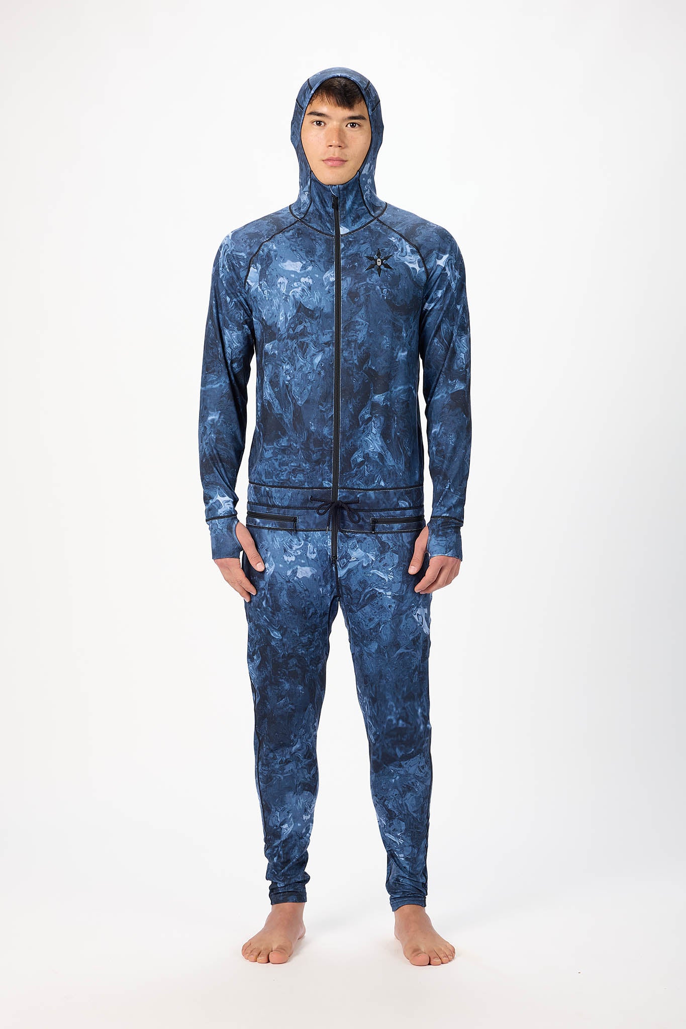 A man wearing a blue swirl patterned onesie with a hooded with a zipper and jogger pants, standing against a plain white studio background.
