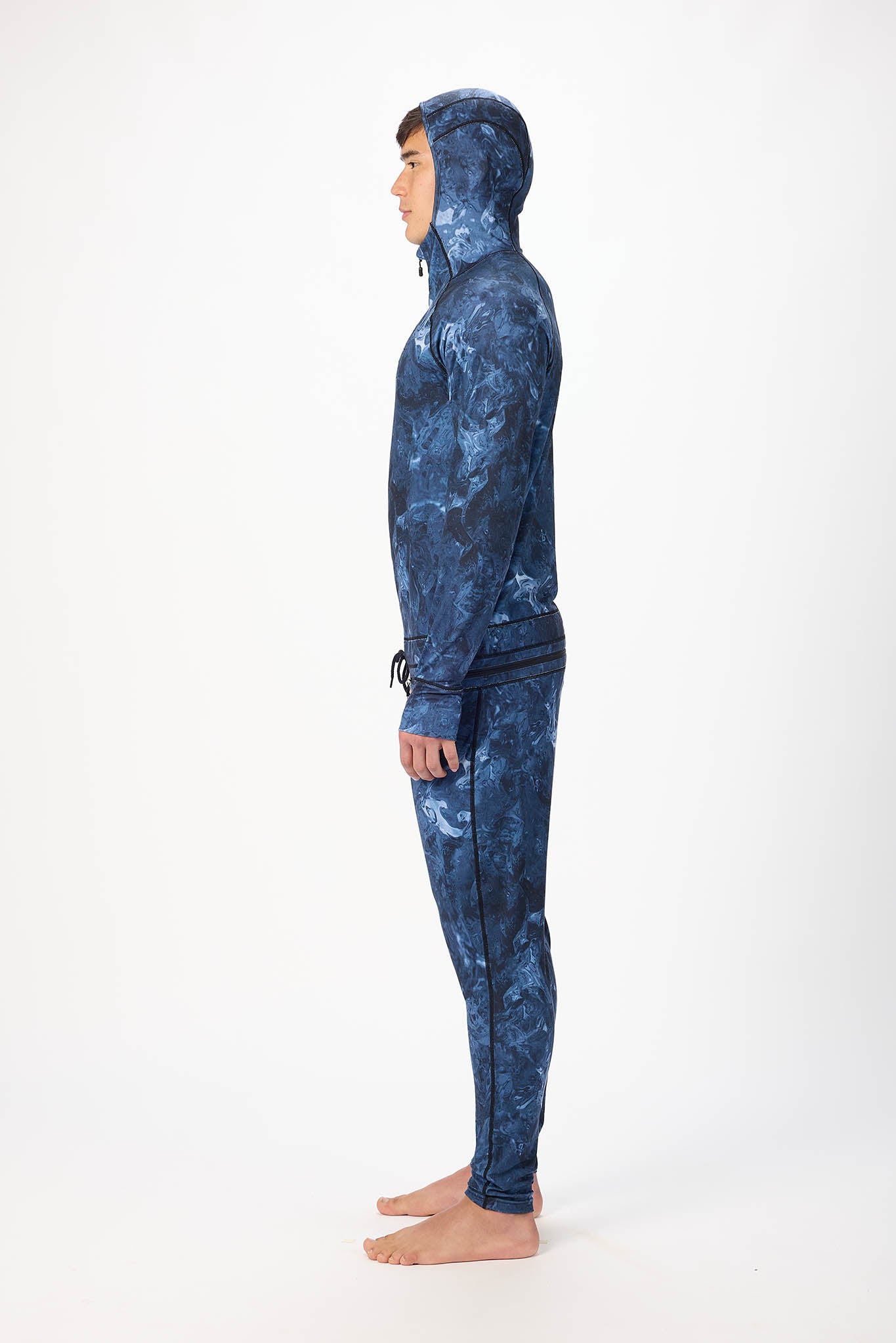 in side view A man wearing a blue swirl patterned onesie with a hooded with a zipper and jogger pants, standing against a plain white studio background.