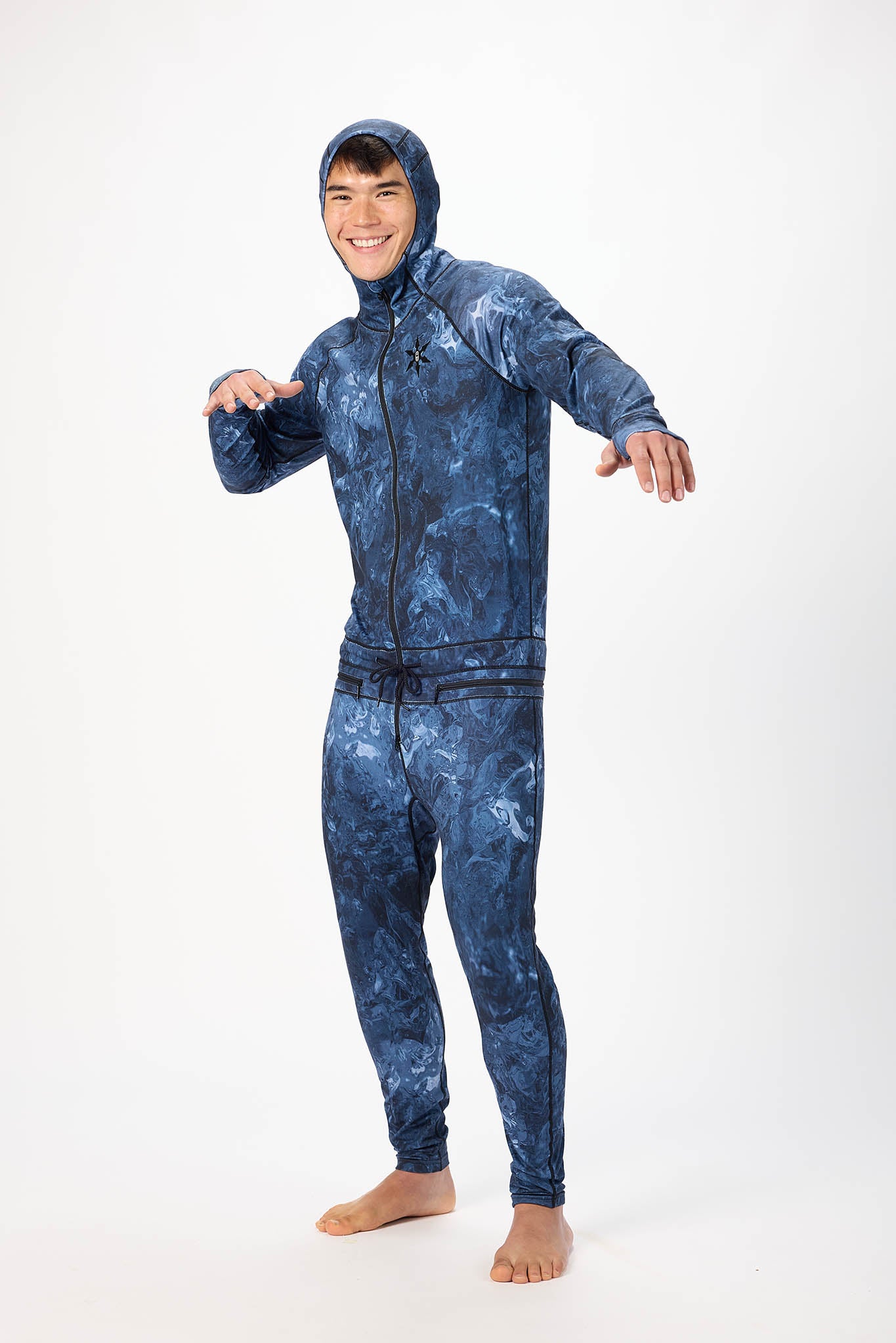A man wearing a blue swirl patterned onesie with a hooded with a zipper and jogger pants, standing against a plain white studio background.