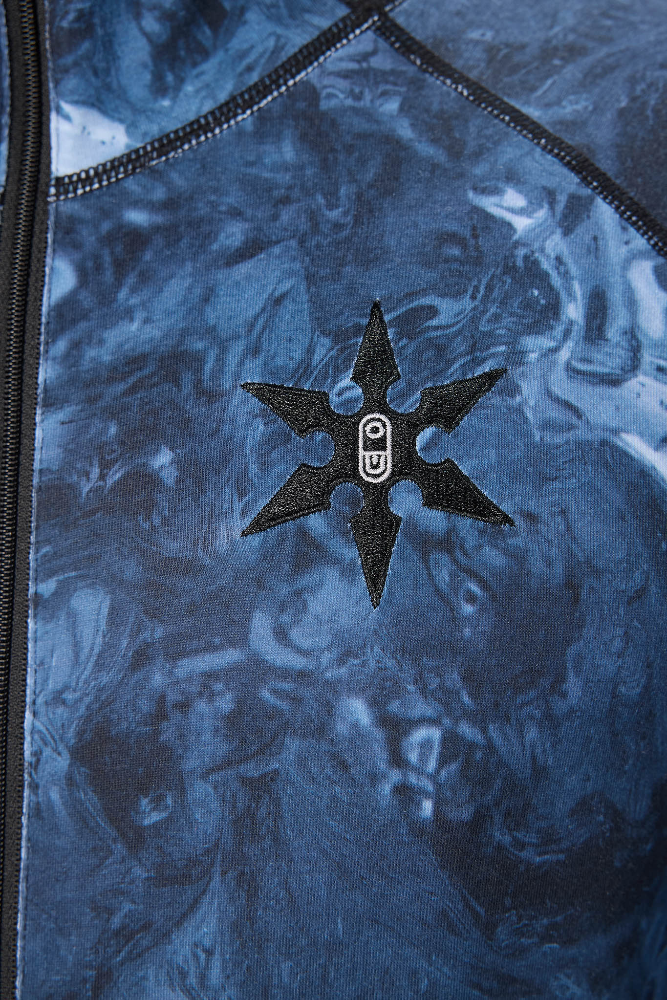 Close-up of a blue marbled fabric garment with a black six-pointed star patch featuring a white circular logo in the center, and a black zipper running along the left edge.