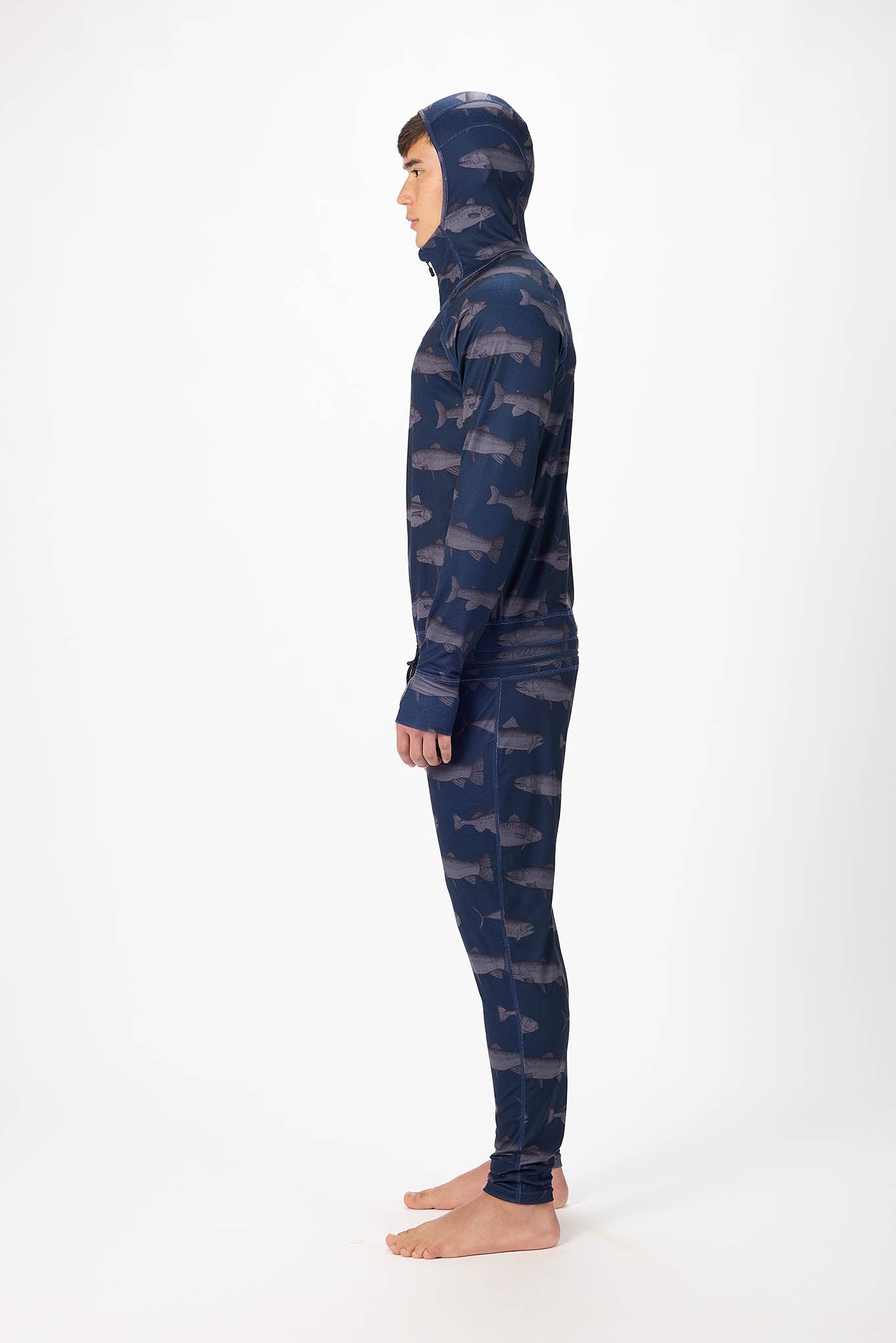 in side profile A man wearing a fish patterned onesie with a hooded with a zipper and jogger pants, standing against a plain white studio background.