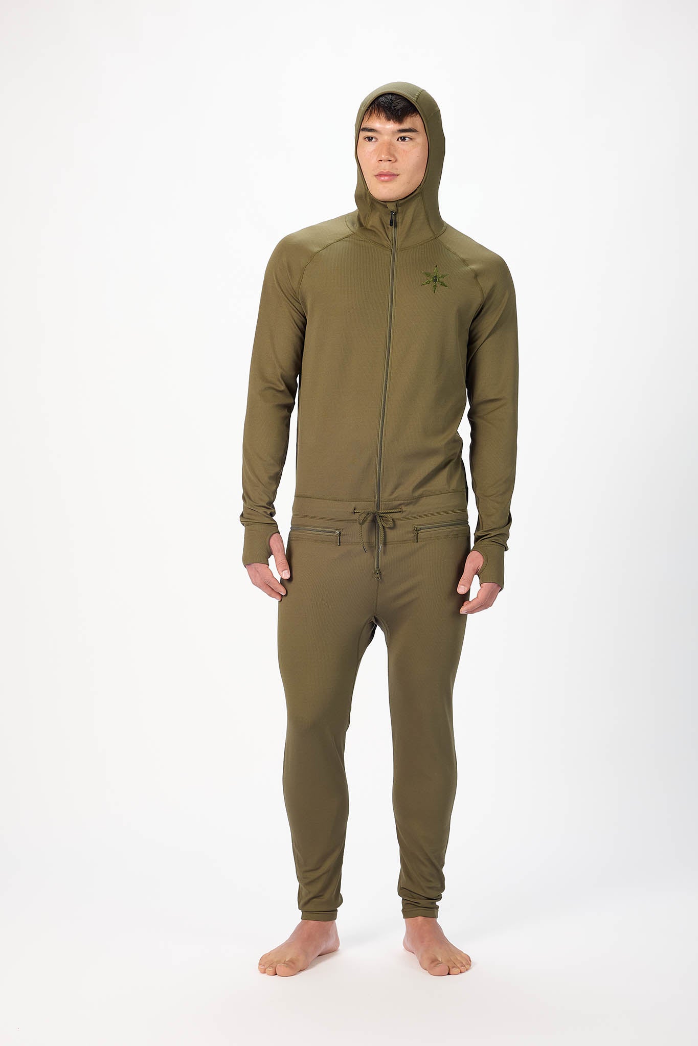 A man wearing a Olive onesie with a hooded with a zipper and jogger pants, standing against a plain white studio background.