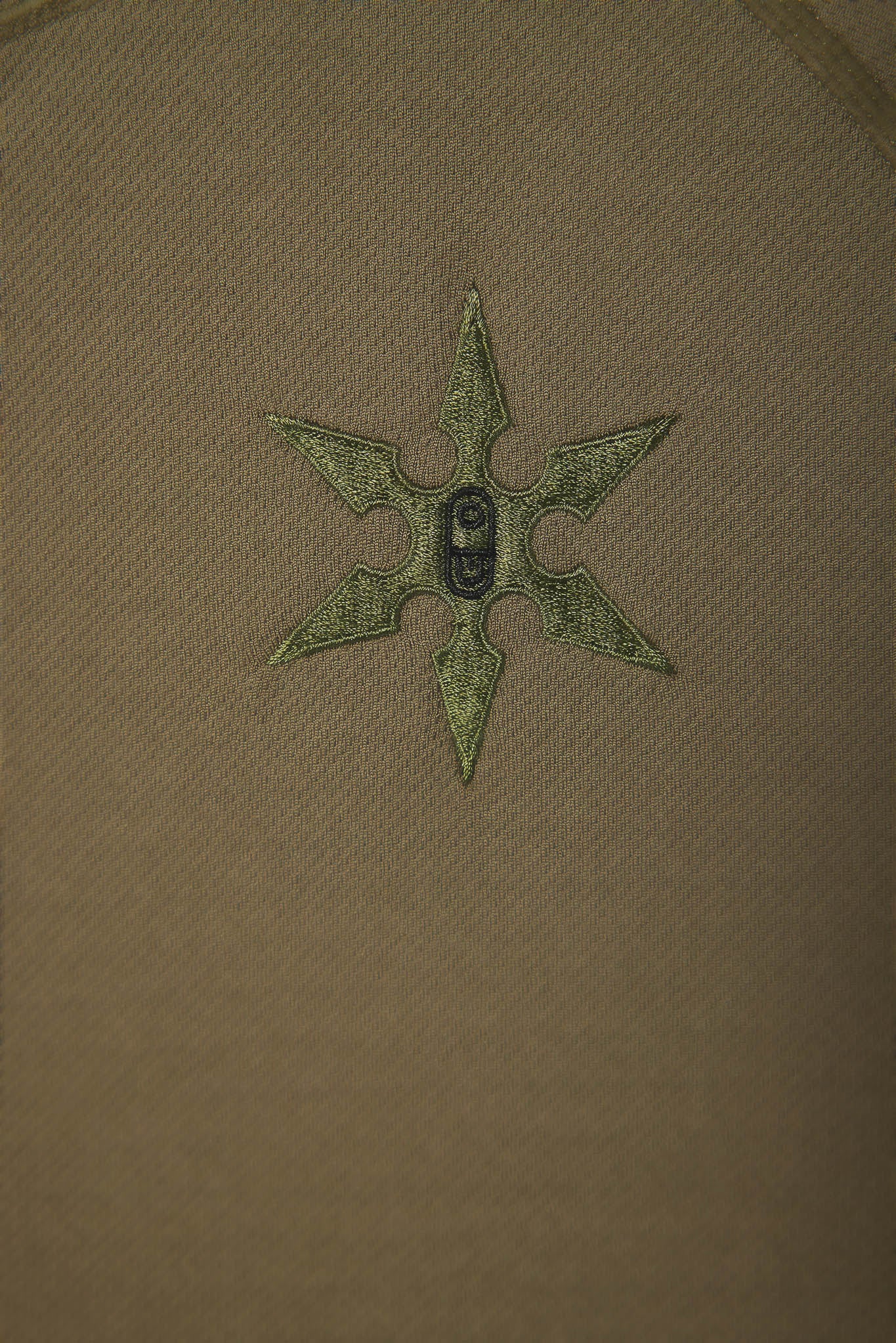 A close-up of a tan or khaki fabric surface with a small embroidered six-pointed star or compass-like motif in olive green and a dark center.