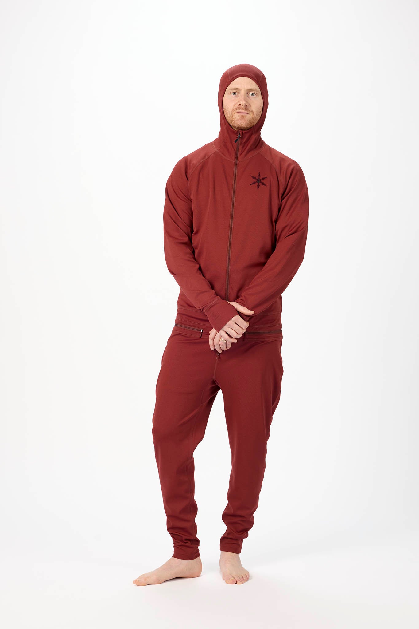 A man wearing a matching burgundy onesie with a hooded zip-up jacket and jogger pants, standing against a plain white studio background.