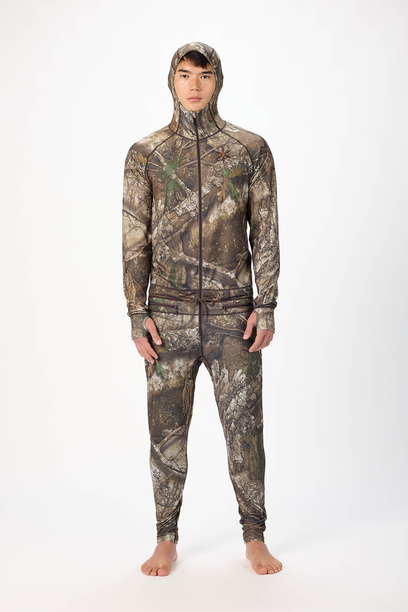 A man wearing a realtree camouflage onesie with a hooded zip-up jacket and jogger pants, standing against a plain white studio background.