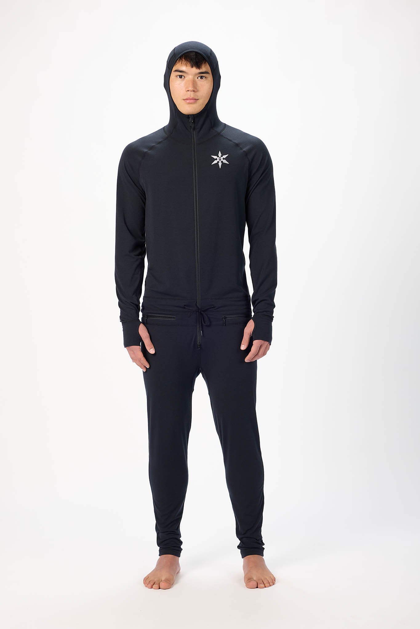 A man wearing a black onesie with a hooded zip-up jacket and jogger pants, standing against a plain white studio background.