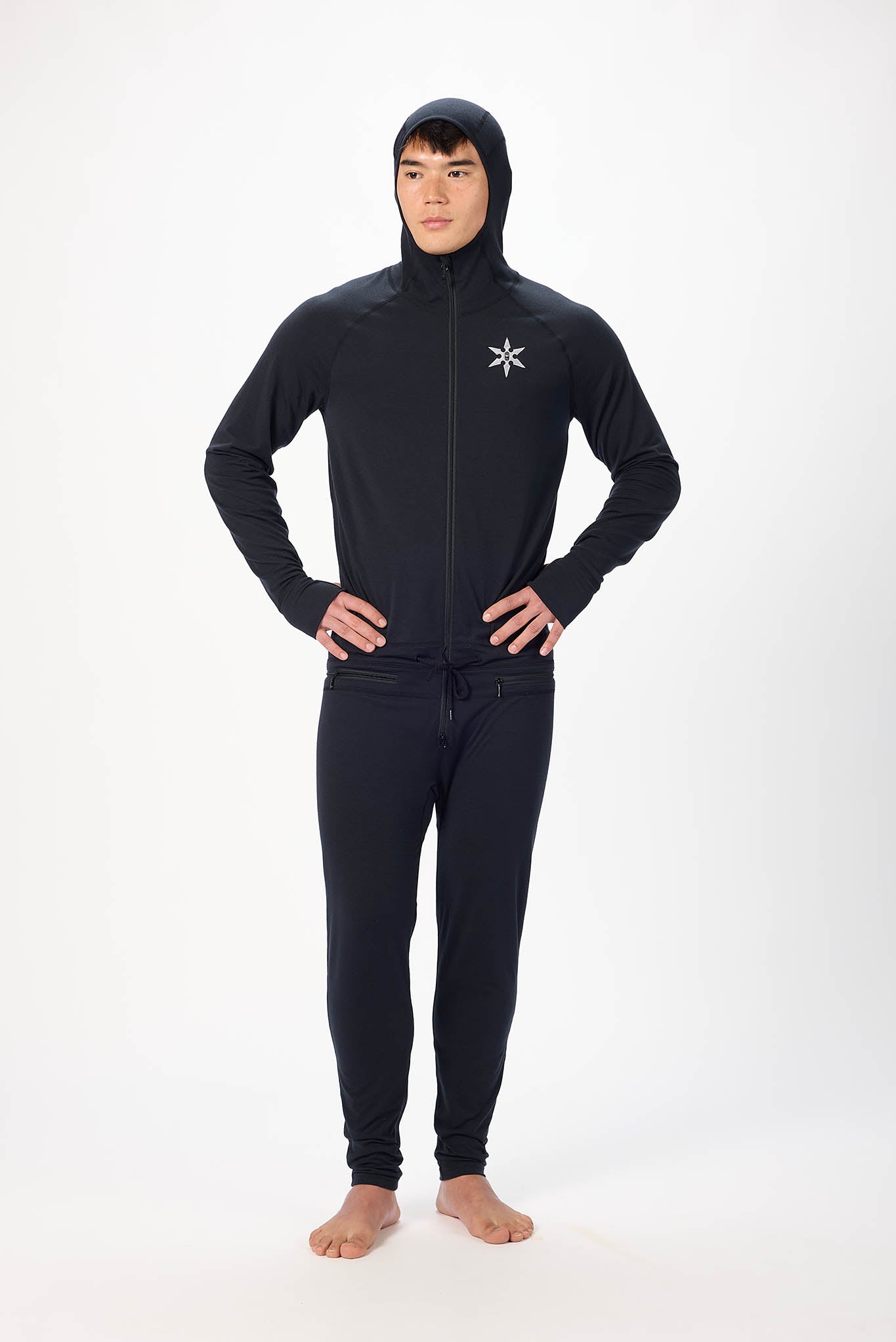 A man wearing a black onesie with a hooded zip-up jacket and jogger pants, standing against a plain white studio background.