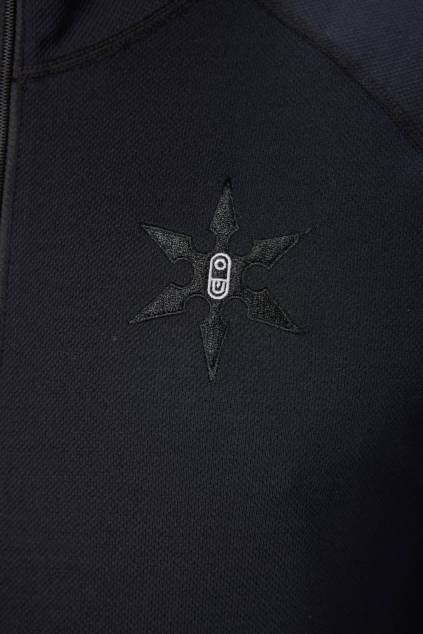 Close-up of a black zippered garment featuring a tonal black embroidered six-pointed star with a small white circular logo in the center.