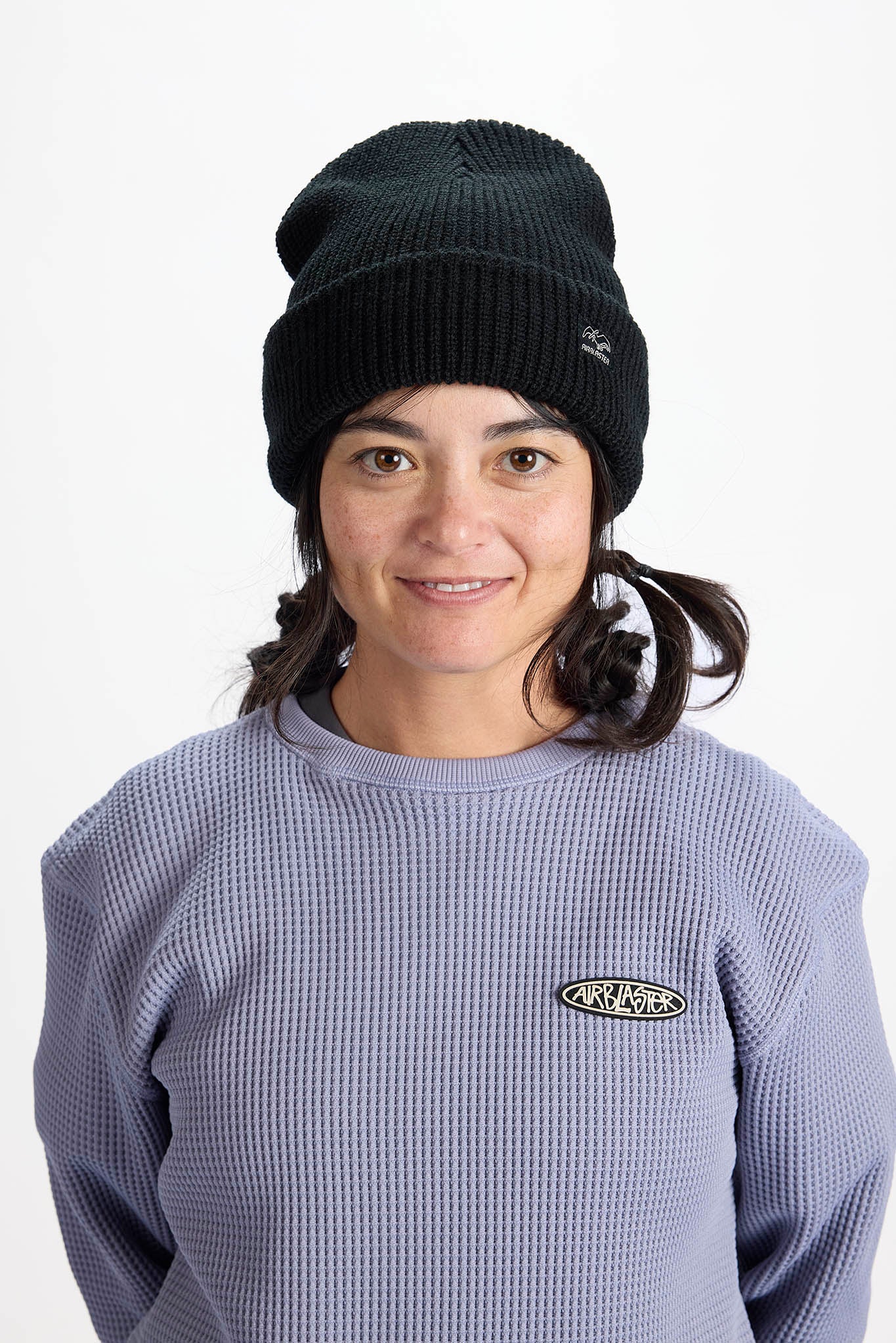 A woman wearing a black ribbed knit beanie and a lavender waffle-knit sweatshirt with a small oval logo on the chest, photographed against a plain light gray background.