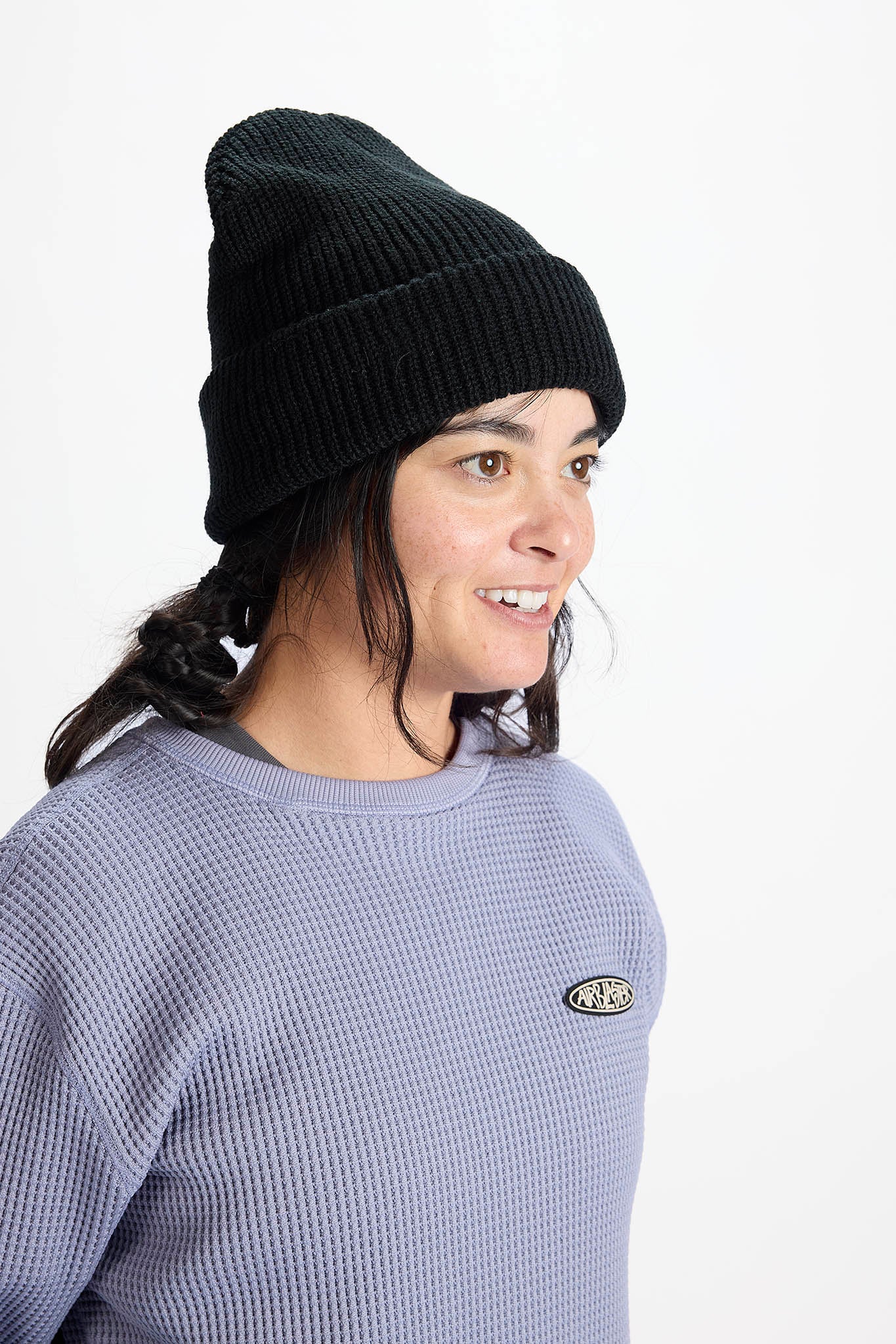 A woman wearing a black ribbed knit beanie and a textured lavender crewneck sweatshirt with a small oval logo patch on the chest, photographed against a plain white studio background.